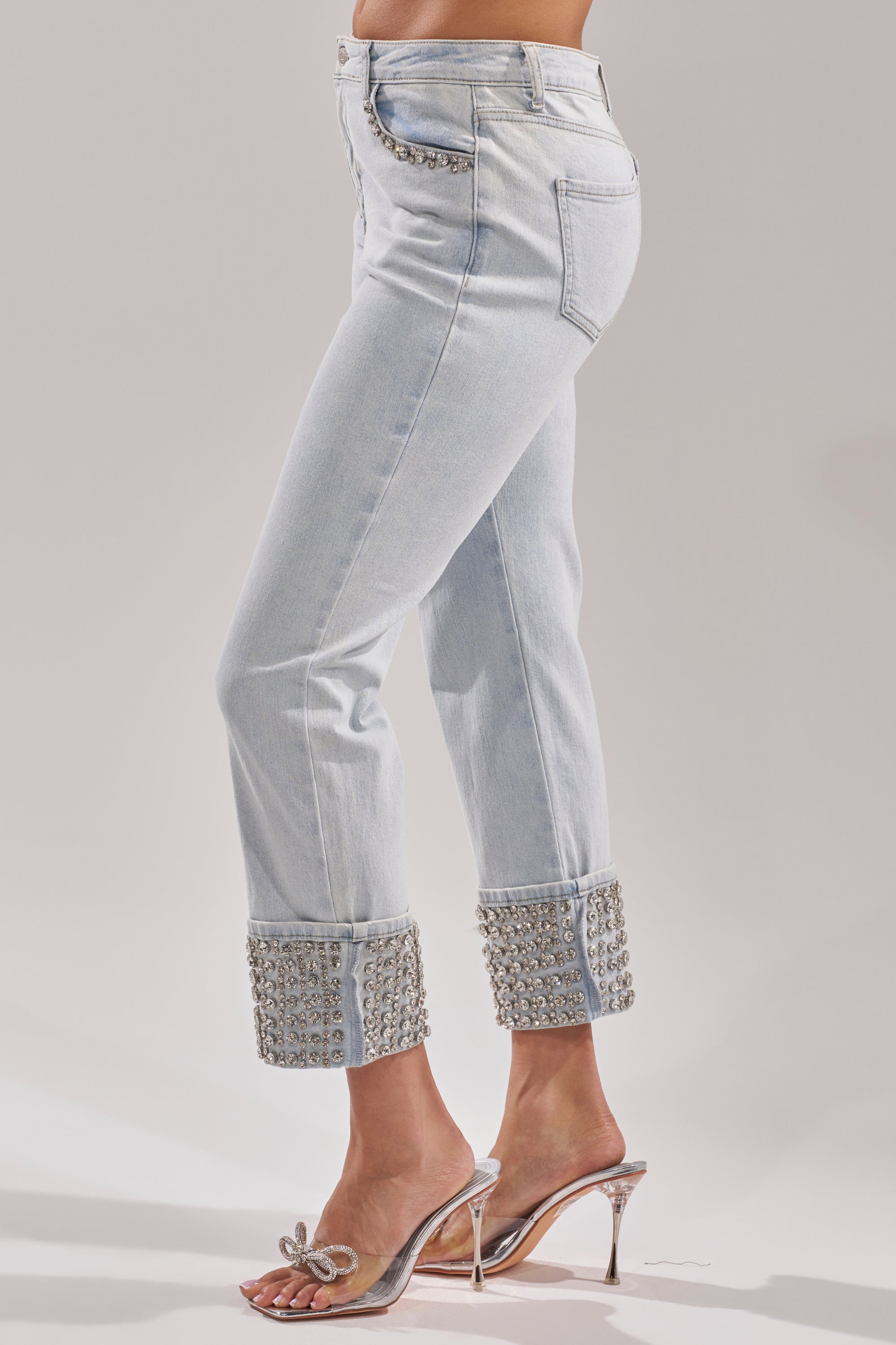 A person wears the HANNI RHINESTONE DENIM CROPPED PANT with large embellished cuffs, paired with clear high-heeled sandals with a bow detail. Only the lower body is visible. 3
