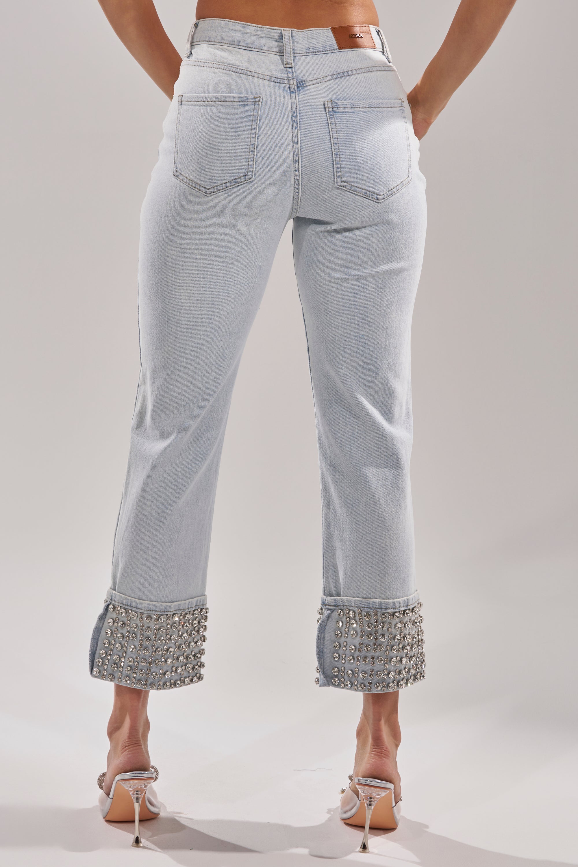 A person faces away from the camera wearing the HANNI RHINESTONE DENIM CROPPED PANT, high-rise jeans with decorative rhinestones adorning the rolled-up hems, paired with clear high-heeled shoes. 4