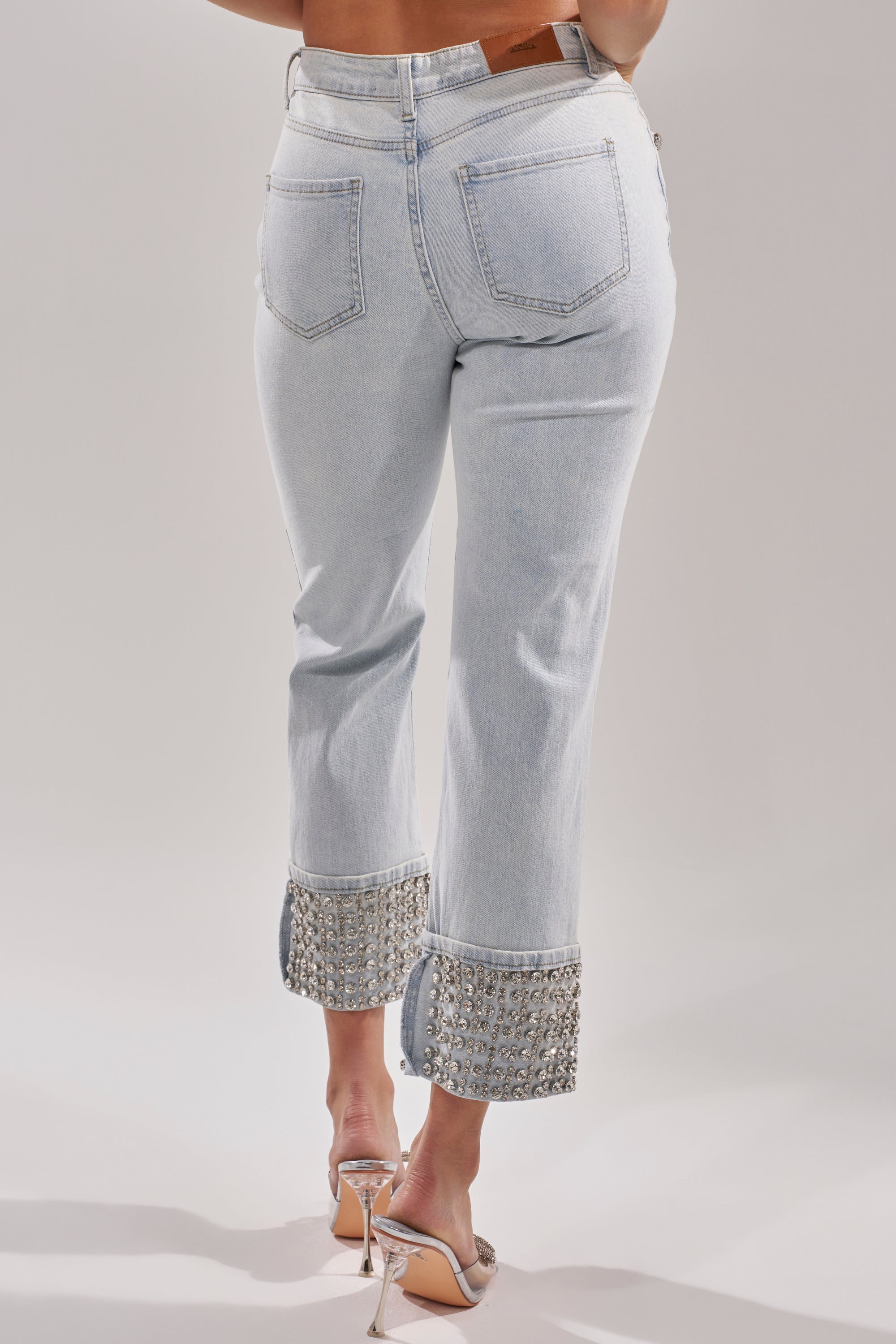A person is seen from the back wearing the HANNI RHINESTONE DENIM CROPPED PANT with embellished cuffs and silver high-heeled sandals, set against a plain background. 5