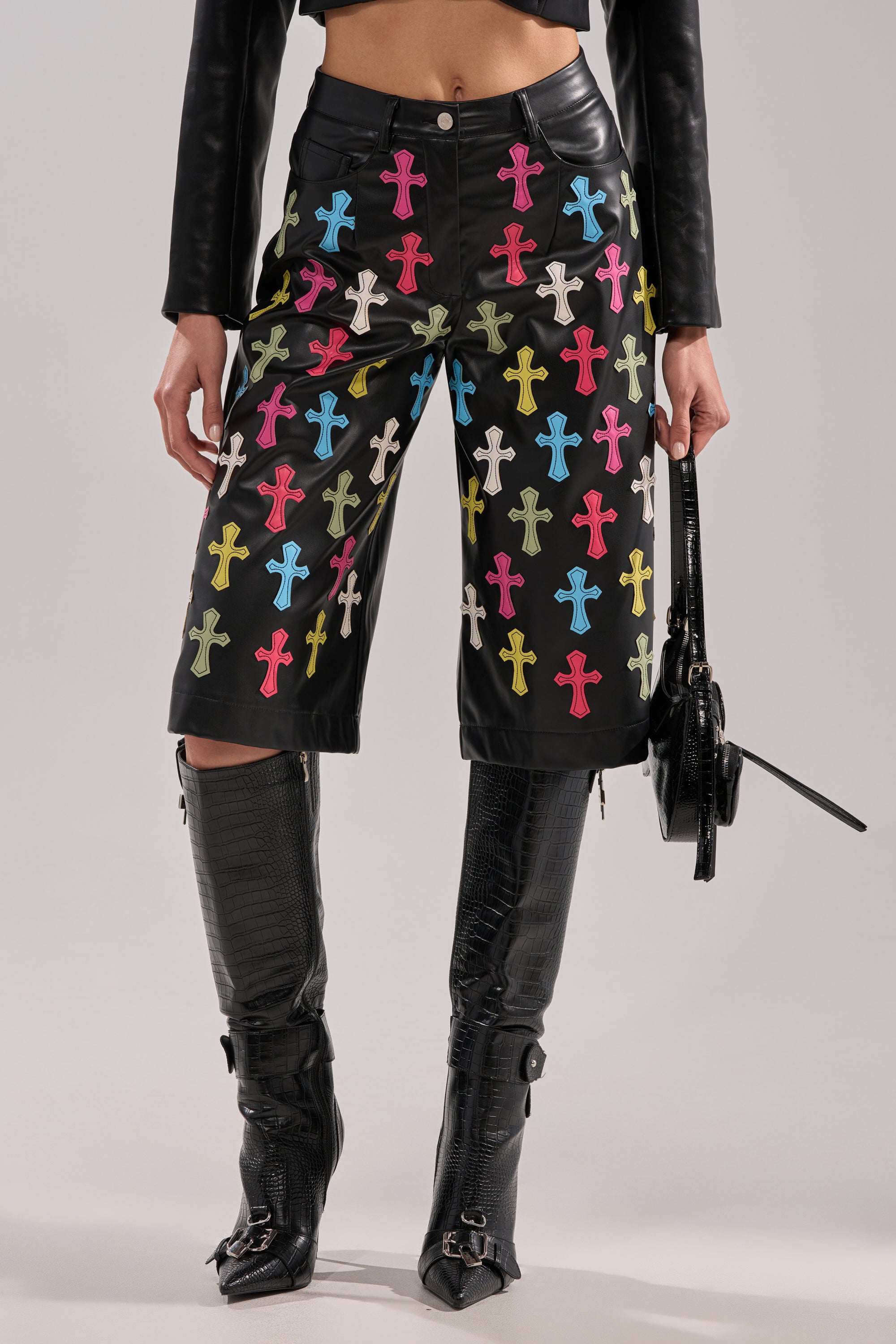 A person wearing the BLESSED SO HARD BERMUDA SHORT paired with a matching black leather jacket, black buckled knee-high boots, and carrying a small black handbag.