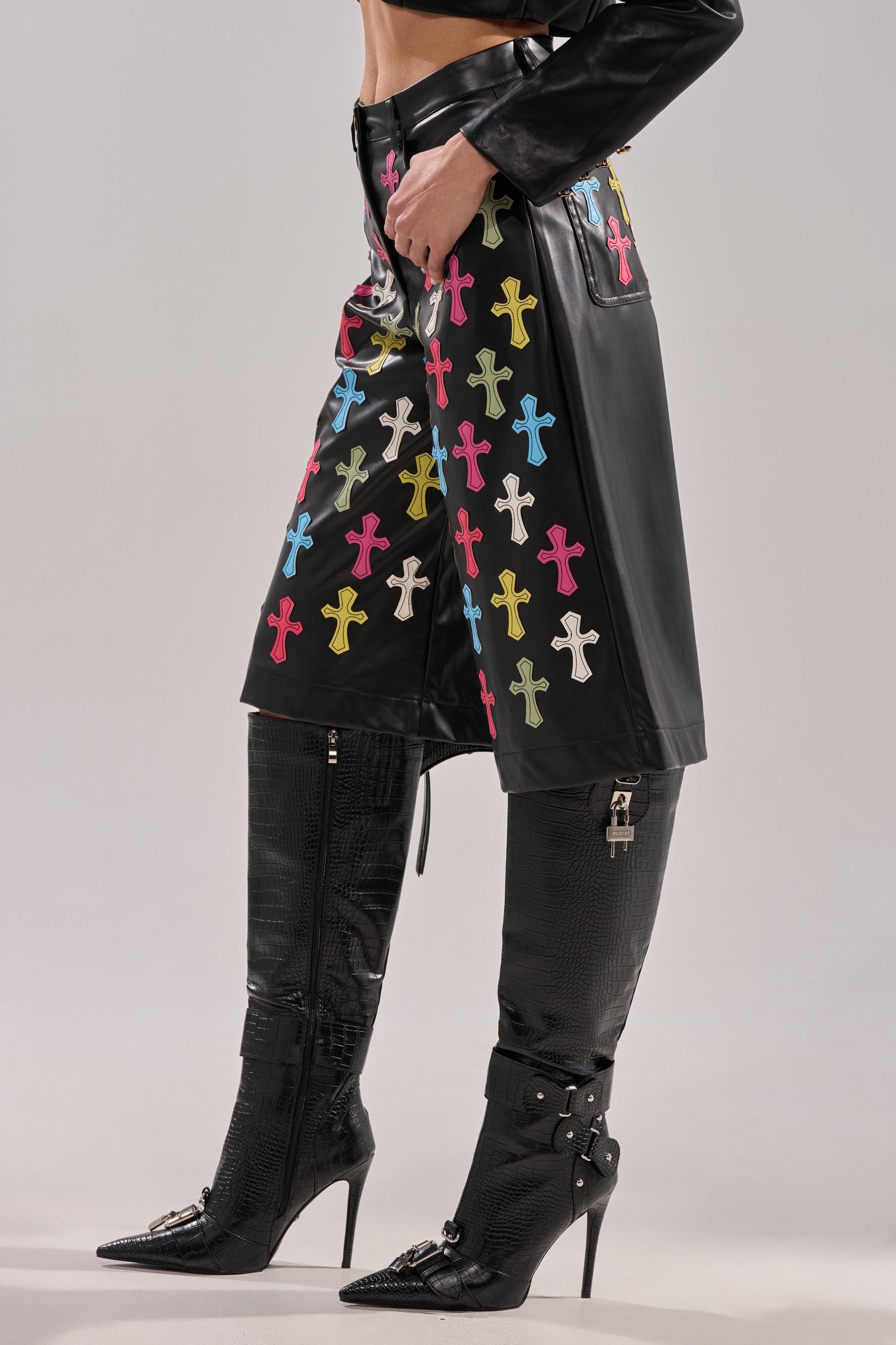 Wearing BLESSED SO HARD BERMUDA SHORT faux leather shorts with multicolored cross appliqués, paired with tall black pointed-toe textured boots with zippers. Only the lower half of the body is shown.