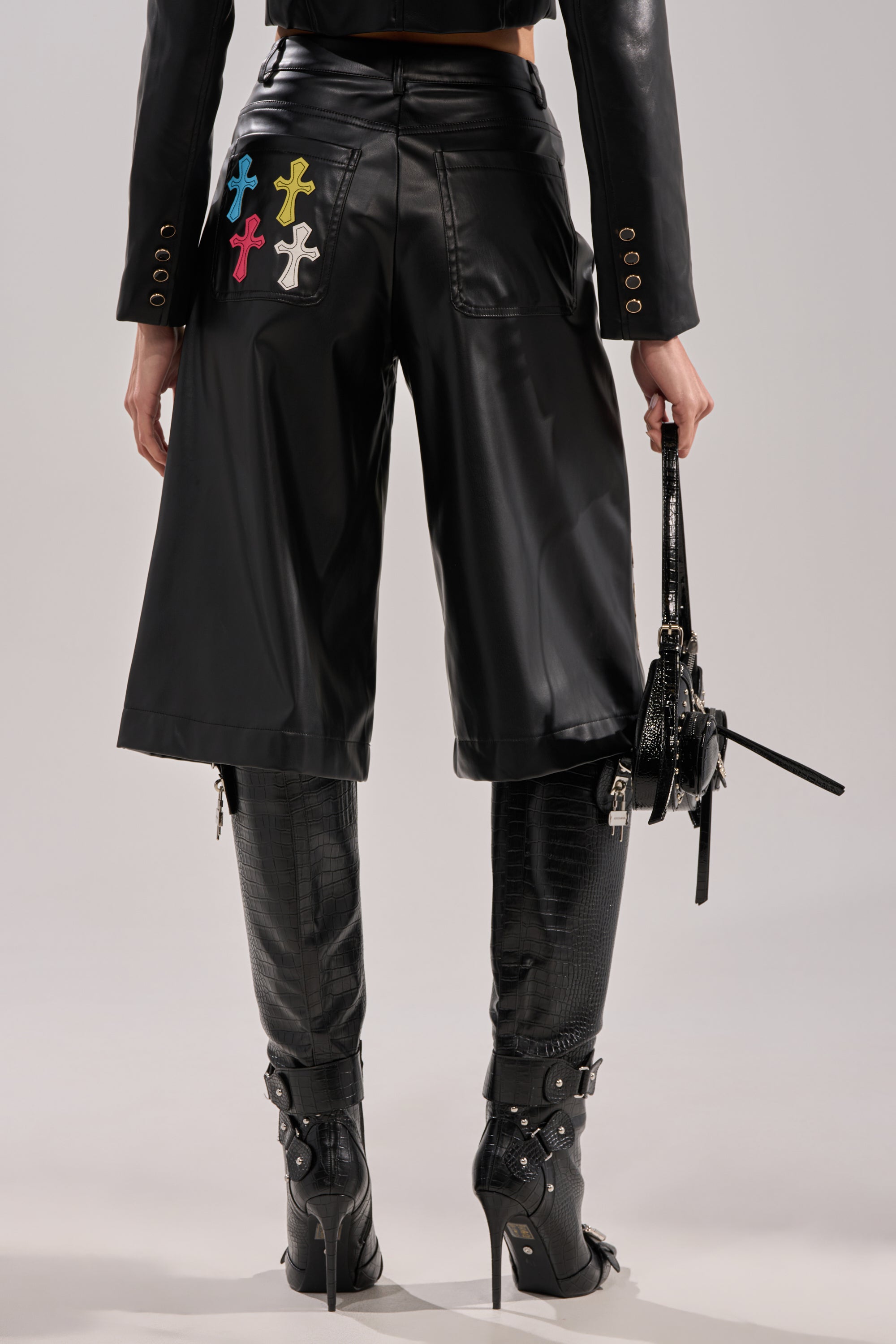 A person wearing BLESSED SO HARD BERMUDA SHORT, black boots with buckles, and holding a small black handbag, viewed from the back.