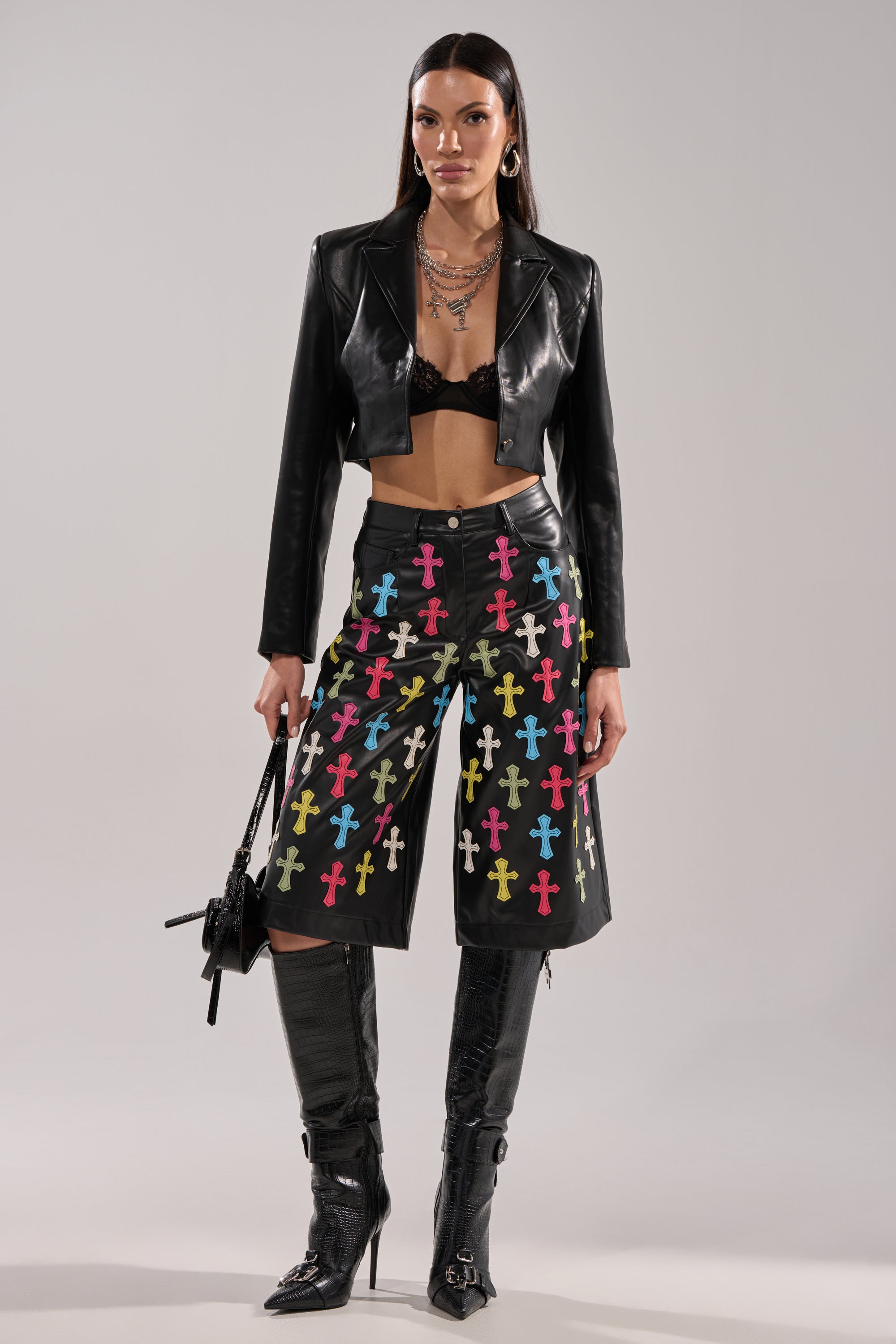A woman wears the BLESSED SO HARD BERMUDA SHORT with multicolored cross appliqués, a black bralette, and a cropped faux leather jacket. She pairs the look with layered necklaces, knee-high boots, and a black handbag.
