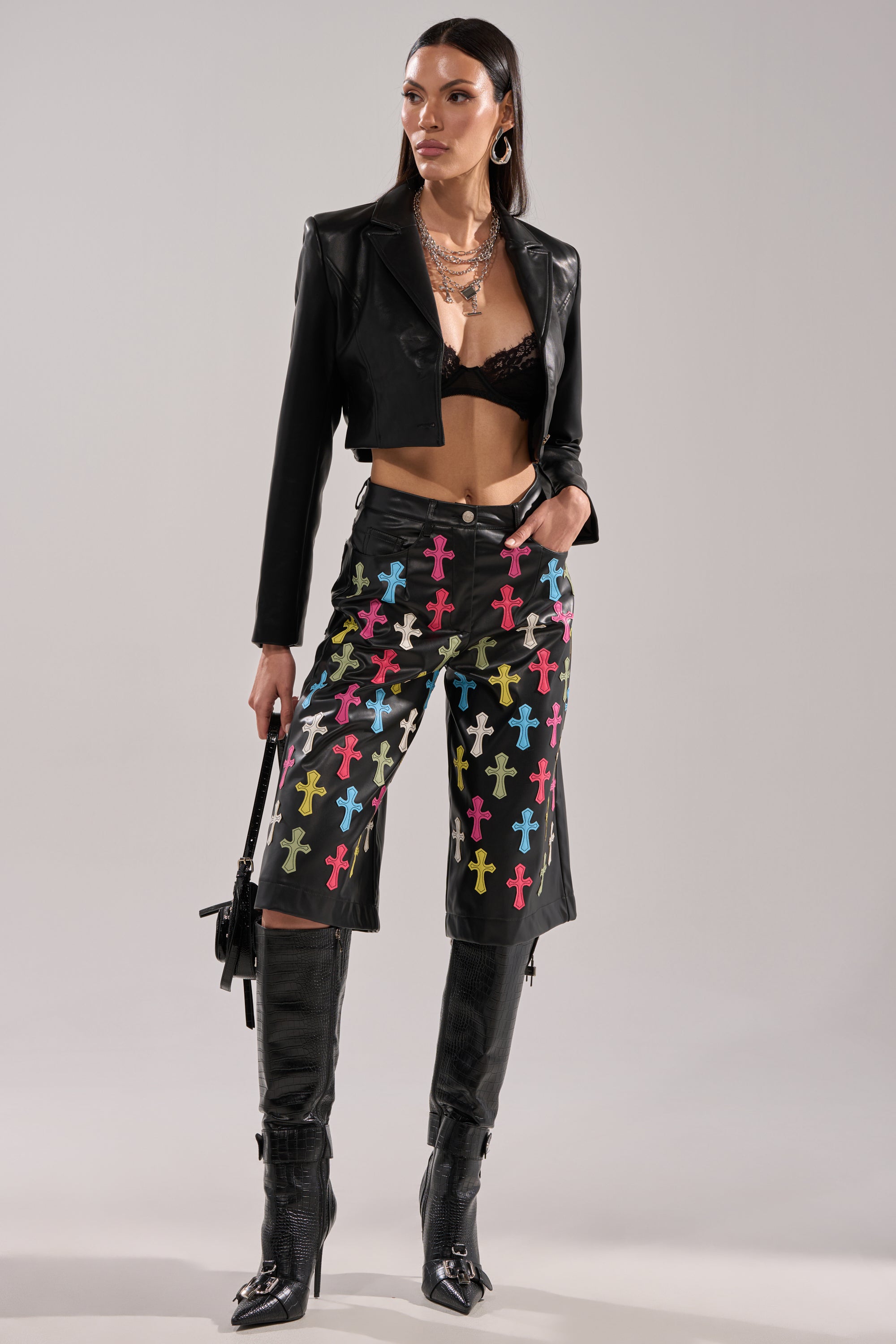 A woman poses in the BLESSED SO HARD BERMUDA SHORT, a pair of faux leather Bermuda shorts with colorful cross patterns, styled with a cropped black leather jacket, lace bralette, tall boots, layered necklaces, hoop earrings, and a small black bag.
