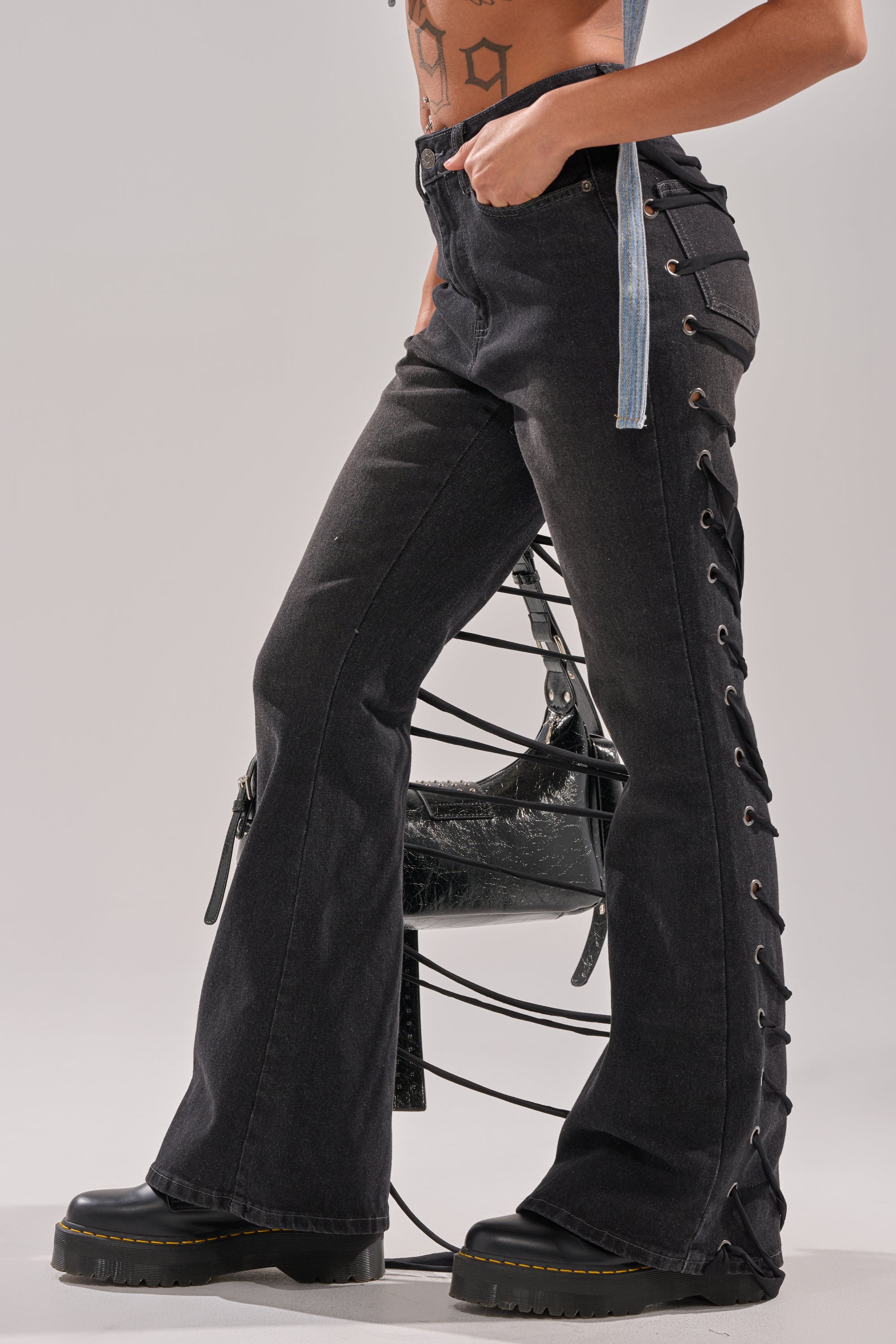 Wearing TRY ME FLARE DENIM with lace-up straps and black platform shoes, a person stands by a modern metal chair. Only their lower torso and legs are shown, emphasizing tattoos and denim strap details on their top. 2