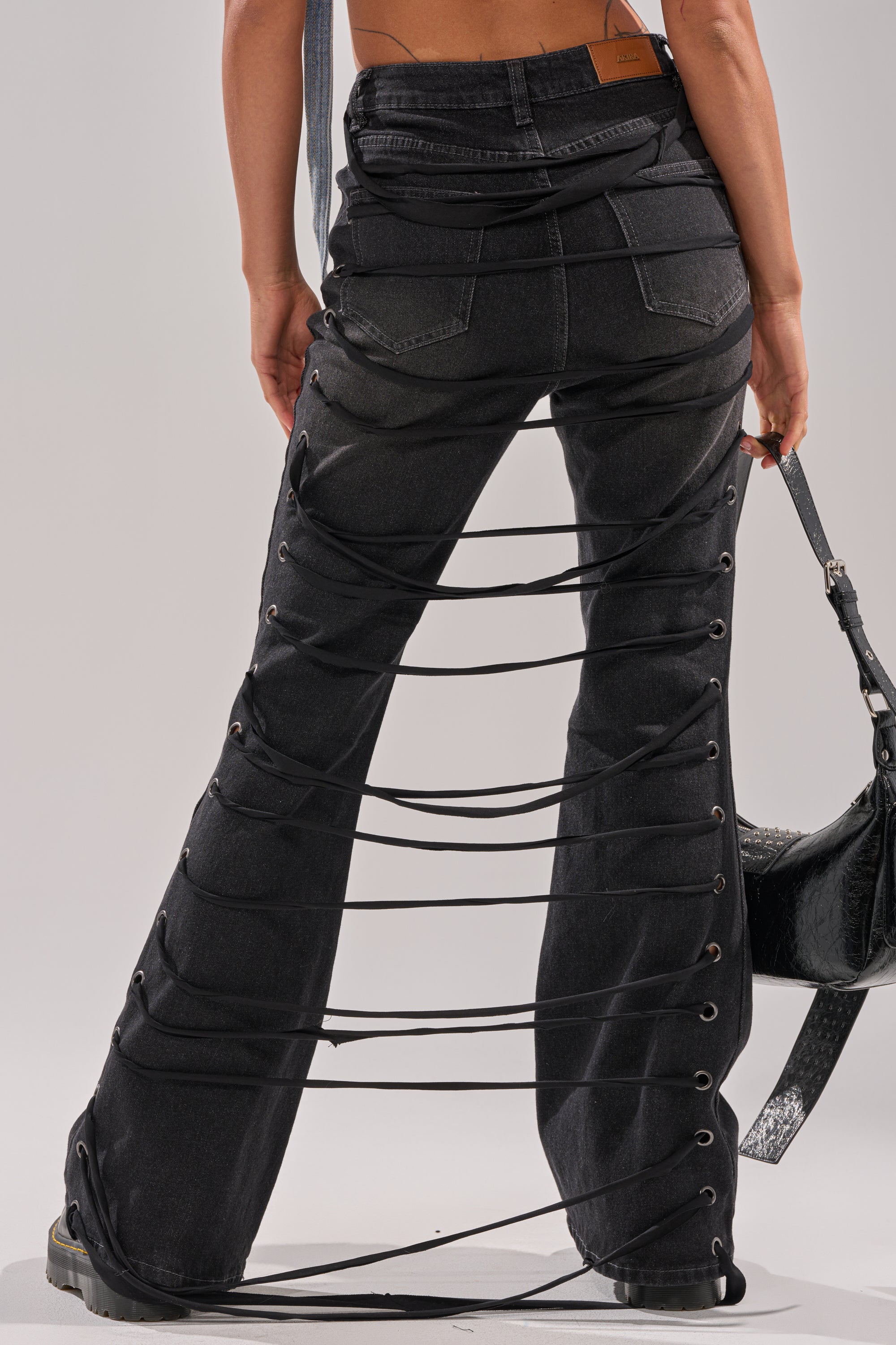 A person wearing TRY ME FLARE DENIM, featuring unique lace-up straps and open sides, holds a black handbag. The photo, taken from behind, highlights the striking crisscross design of the pants. 3