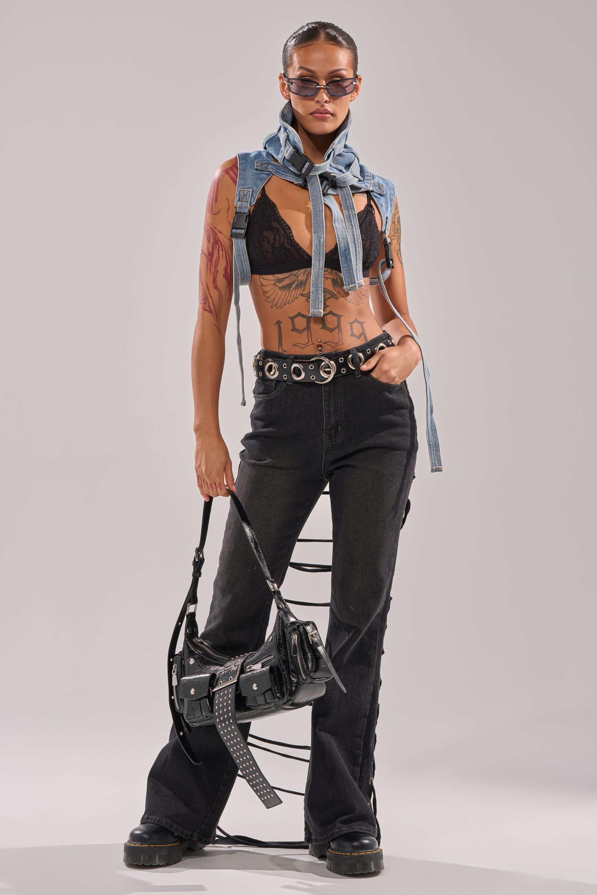 Wearing the TRY ME FLARE DENIM high-rise black jeans with lace-up straps, a black lace bralette, sunglasses, a sleeveless denim jacket tied around the shoulders, and holding a black handbag, they pose confidently against a plain background. 4