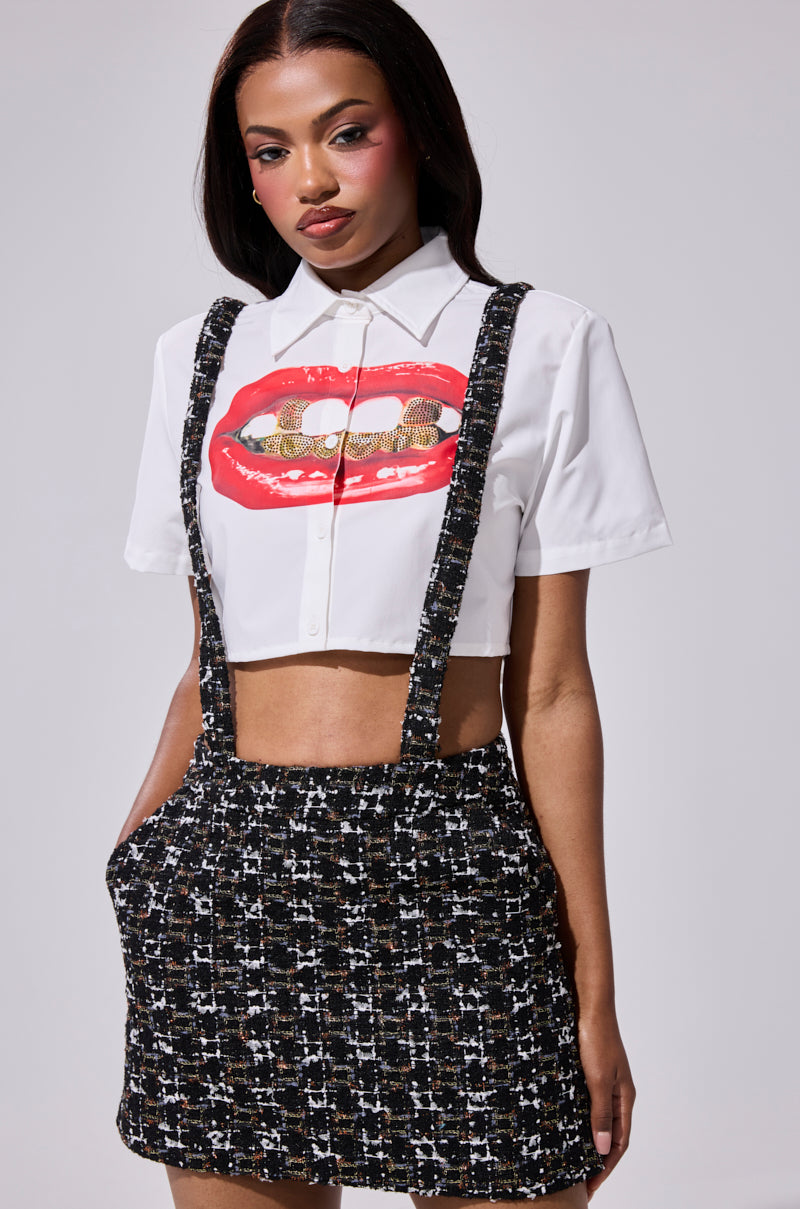 A woman stands with hands in pockets against a plain background, wearing a white cropped shirt with red lips and gold grills graphic, paired with the GIRLY POP TWEED OVERALL SKIRT in a black and white pattern.