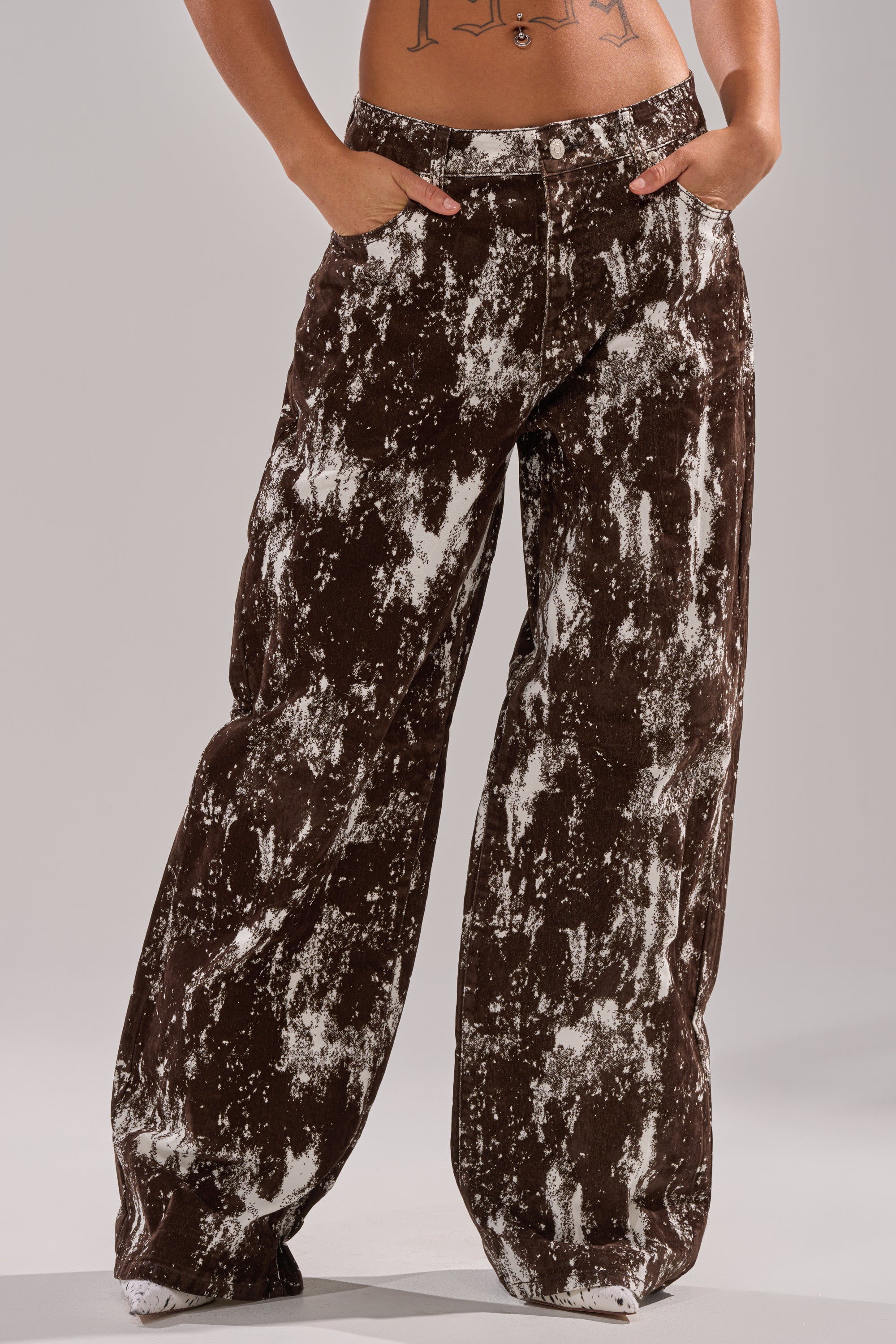 A person wears the CRAZY IN LOVE TROUSER, showcasing its brown wide-leg silhouette with a white splatter pattern, hands in pockets and revealing their midriff and tattoos. 1