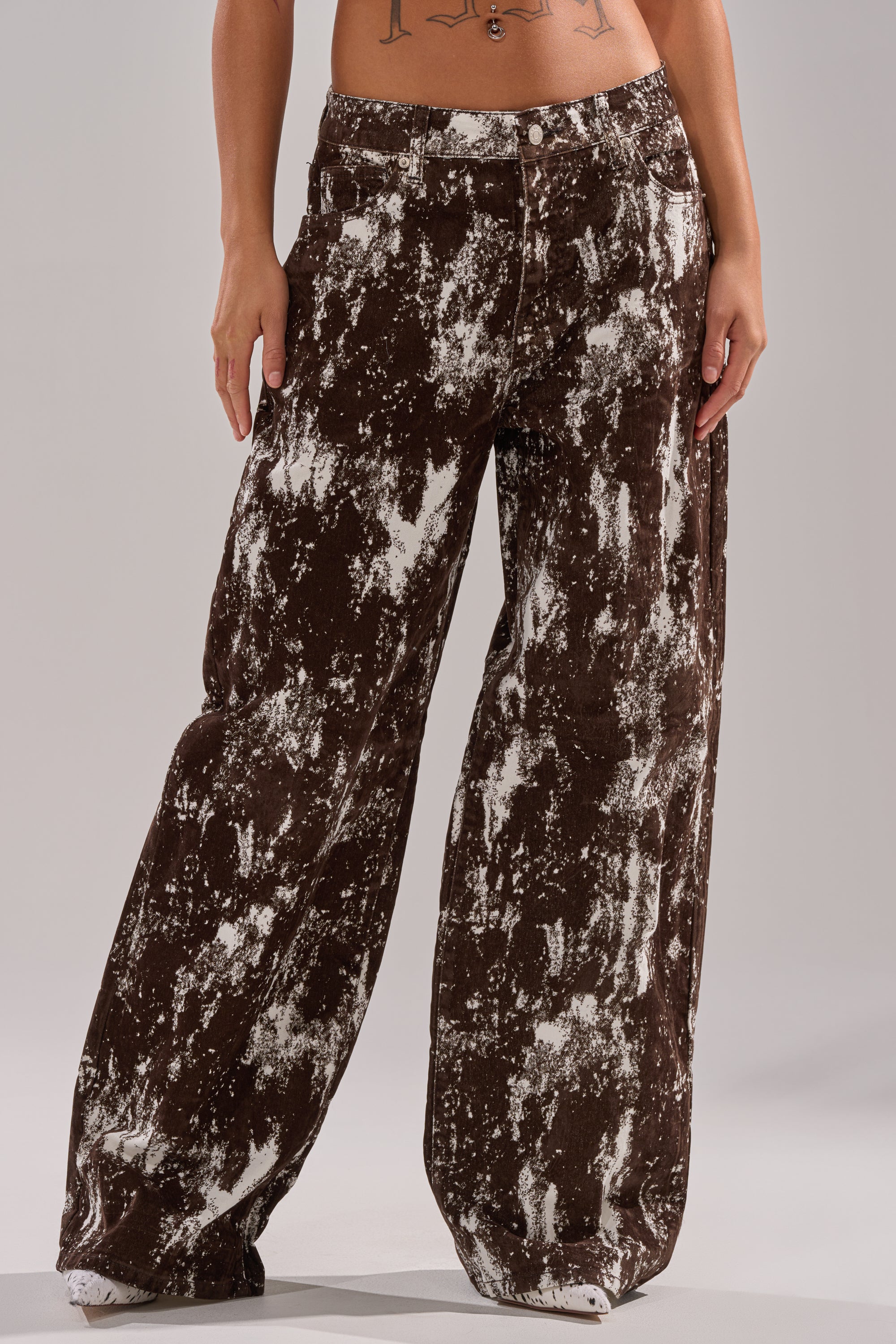 Someone wears the CRAZY IN LOVE TROUSER—a high-waisted, wide-leg, loose-fitting brown pant with a white splatter pattern. Only their lower torso and legs are shown, standing with one hand by their side. 2