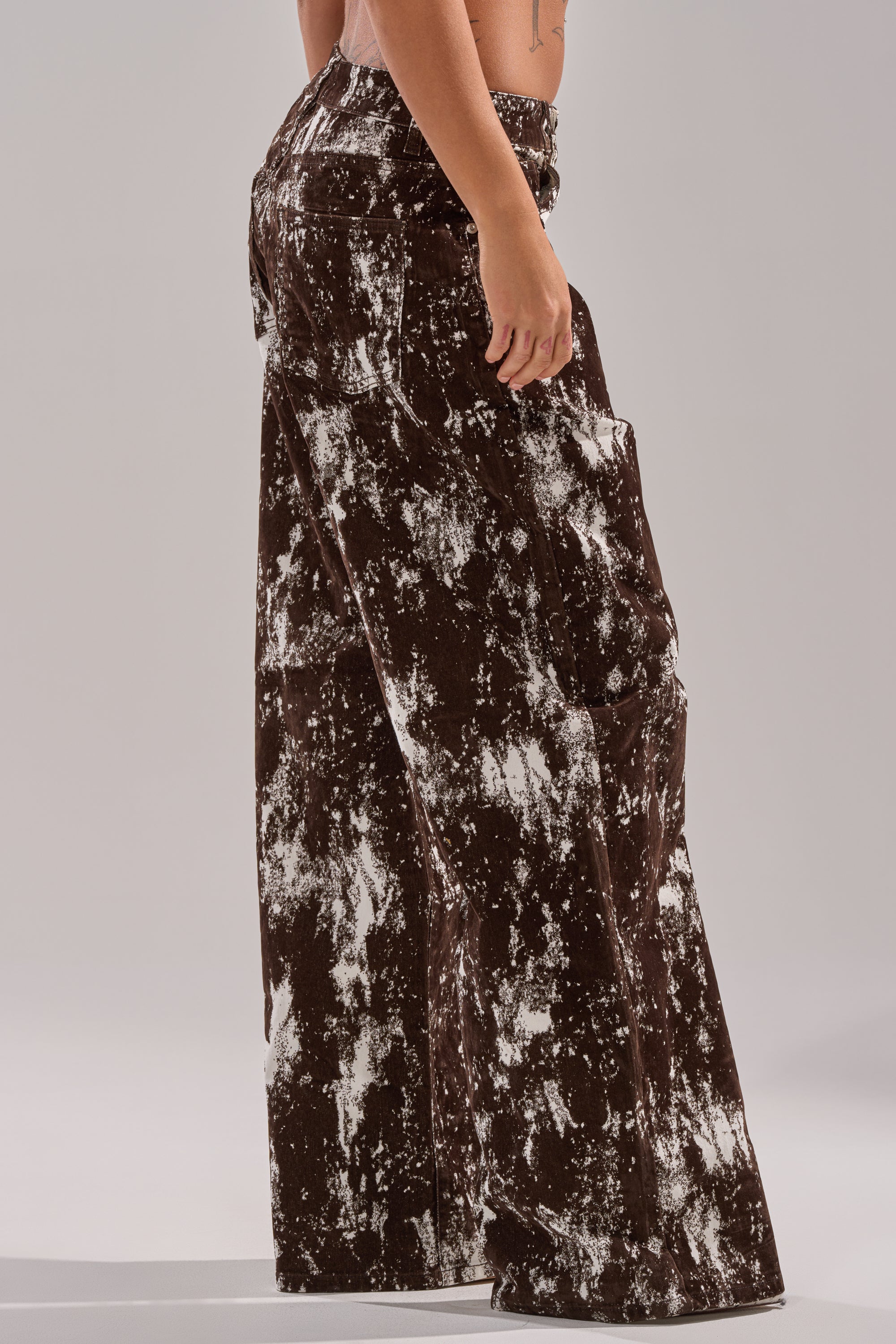 A model showcases the CRAZY IN LOVE TROUSER, featuring a high-waisted, wide-leg silhouette in a brown and white tie-dye design, set against a plain light gray background. 3