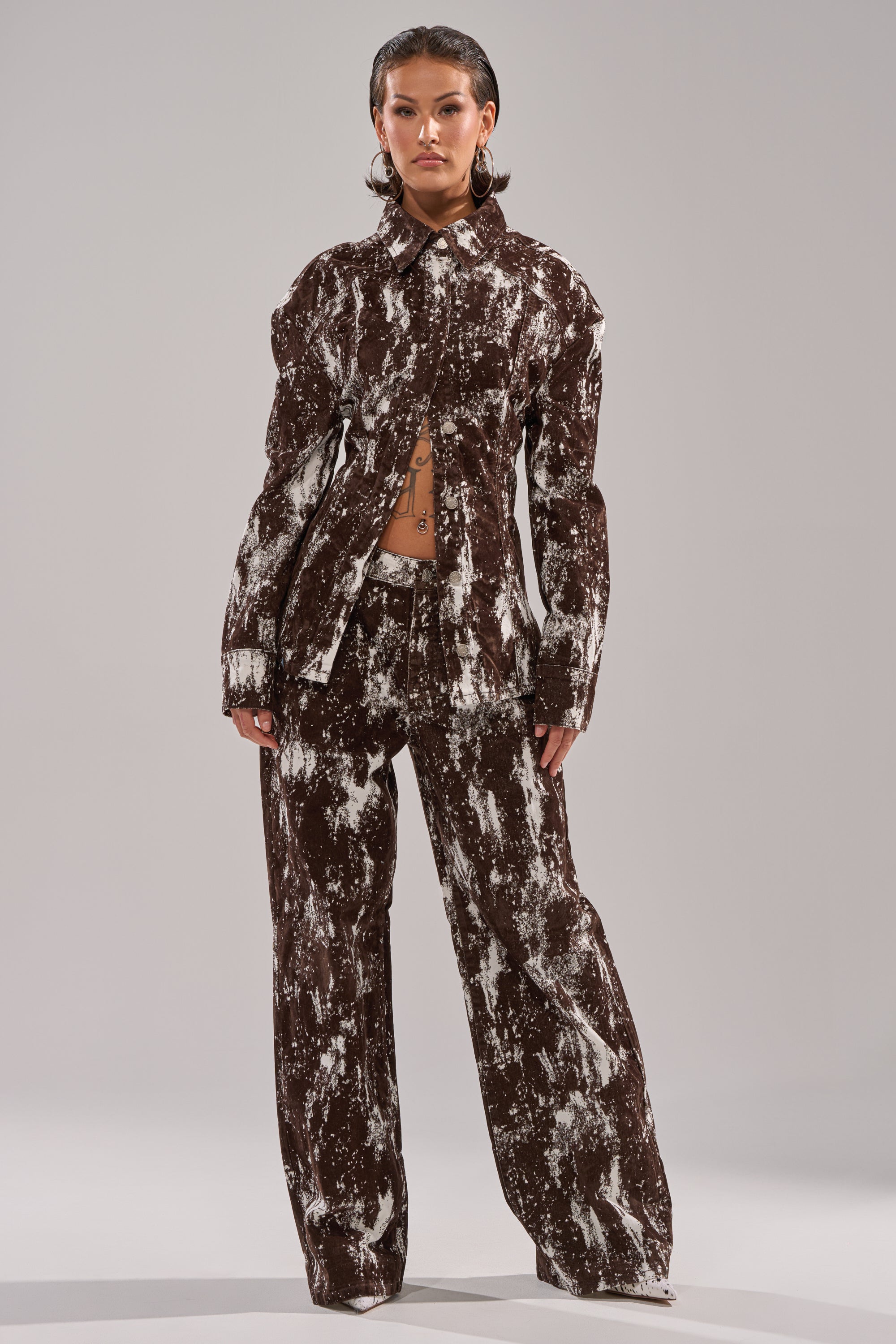 A person stands facing forward in the CRAZY IN LOVE TROUSER suit, featuring a brown and white tie-dye pattern, wide-leg silhouette, and buttoned-up jacket. They have slicked-back hair, visible abdominal tattoos, and a plain light background. 5