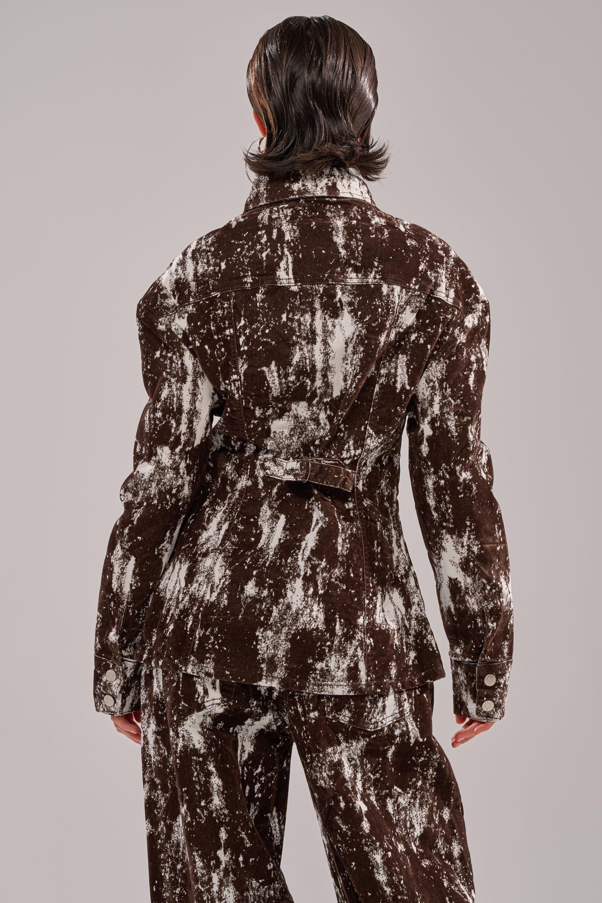A person with short, slicked-back hair faces away, wearing the CRAZY IN LOVE BUTTON DOWN in a brown and white abstract print with matching wide-leg pants. The background is plain and light gray. 3