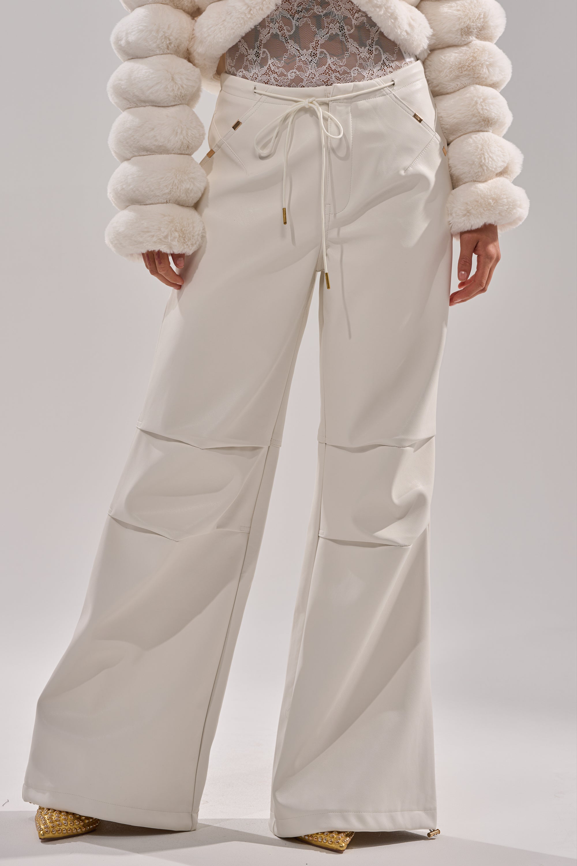 A person wearing the GET WITH IT FAUX LEATHER TROUSER IN WHITE, a white lace top, a textured fur jacket, and gold pointed-toe shoes stands against a plain background. 0