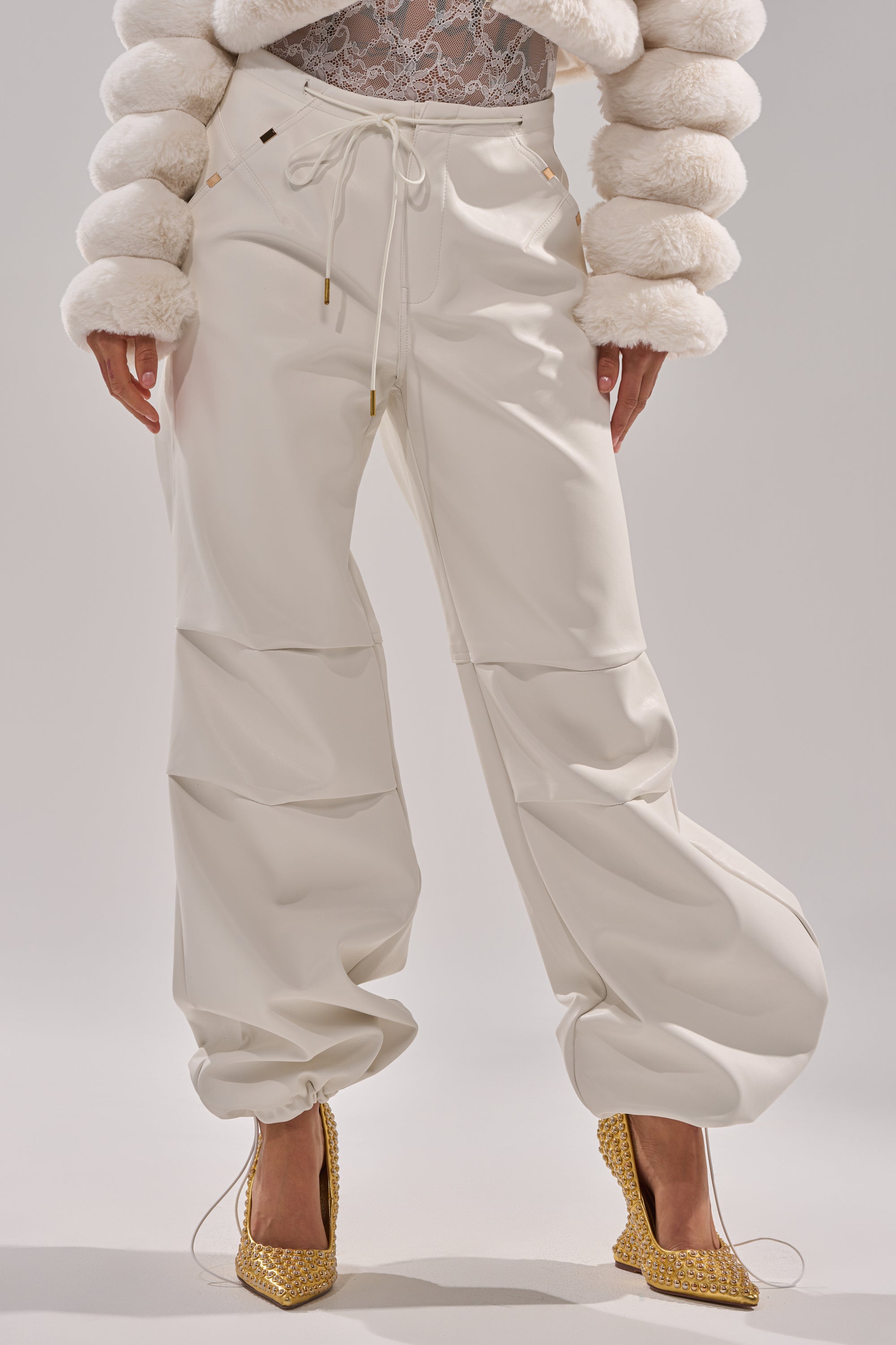 A person stands against a plain light background wearing the GET WITH IT FAUX LEATHER TROUSER IN WHITE, featuring a loose, high-waisted wide-leg fit with drawstrings, paired with a textured white jacket and gold high heels. 1