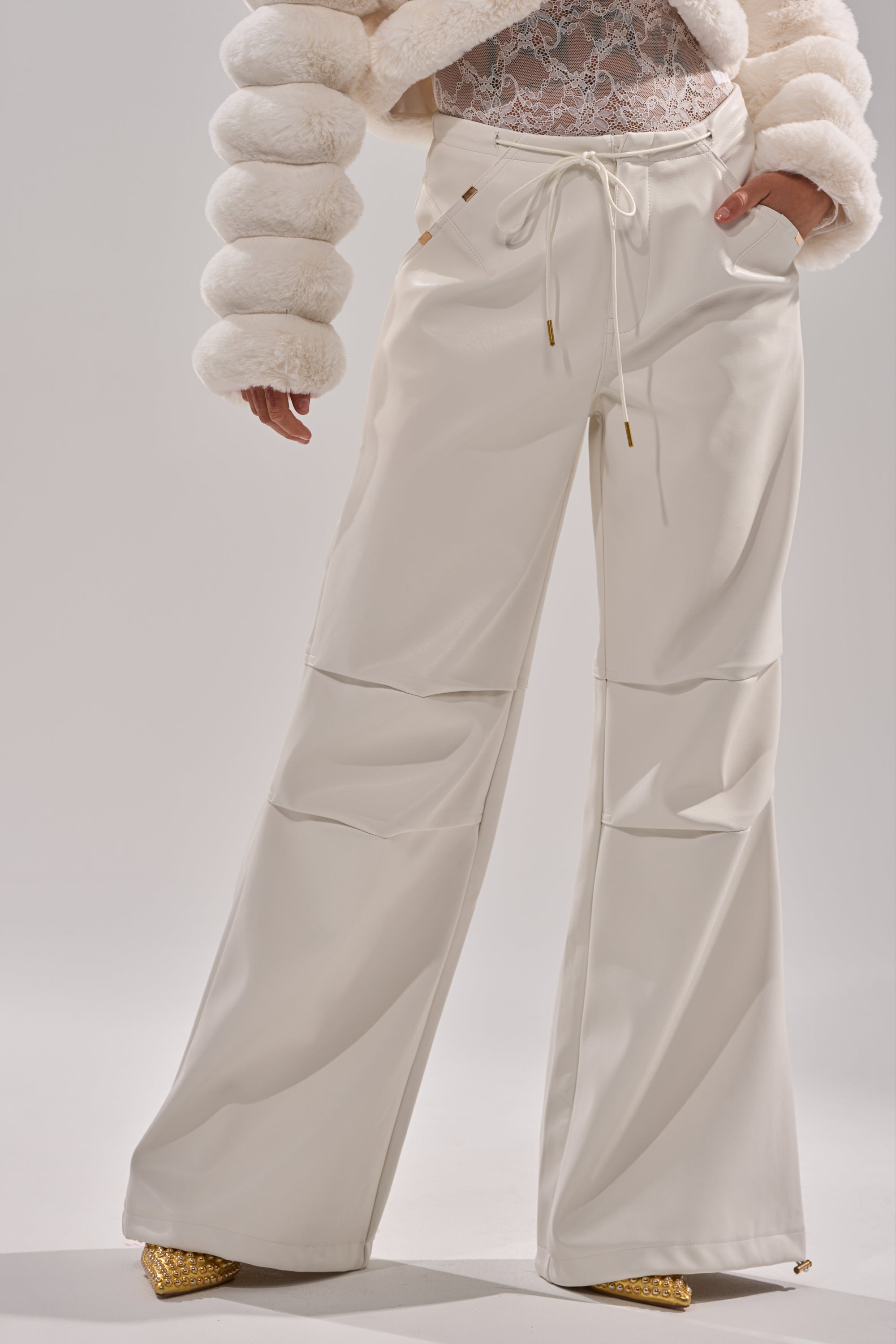 A person styles the GET WITH IT FAUX LEATHER TROUSER IN WHITE—wide-leg pants with drawstrings—paired with a white lace top, textured fur jacket, and gold pointed-toe shoes against a minimal background for an effortlessly chic look. 2