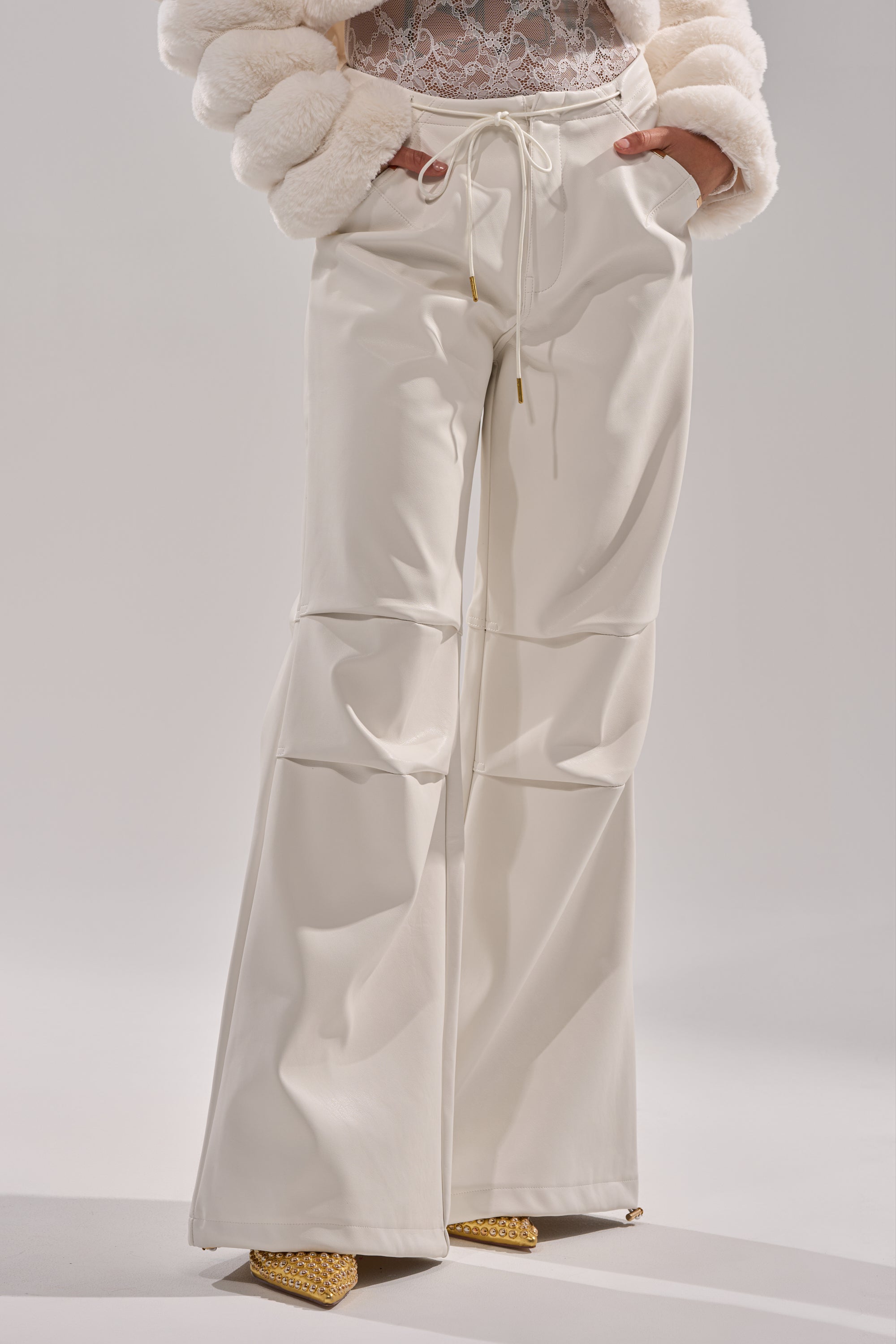 A person wearing GET WITH IT FAUX LEATHER TROUSER IN WHITE, featuring a high-waisted wide-leg fit with drawstrings. The outfit includes a lace top, fur jacket, and yellow pointed-toe shoes. Photo is cropped at the shoulders to showcase the trousers. 3