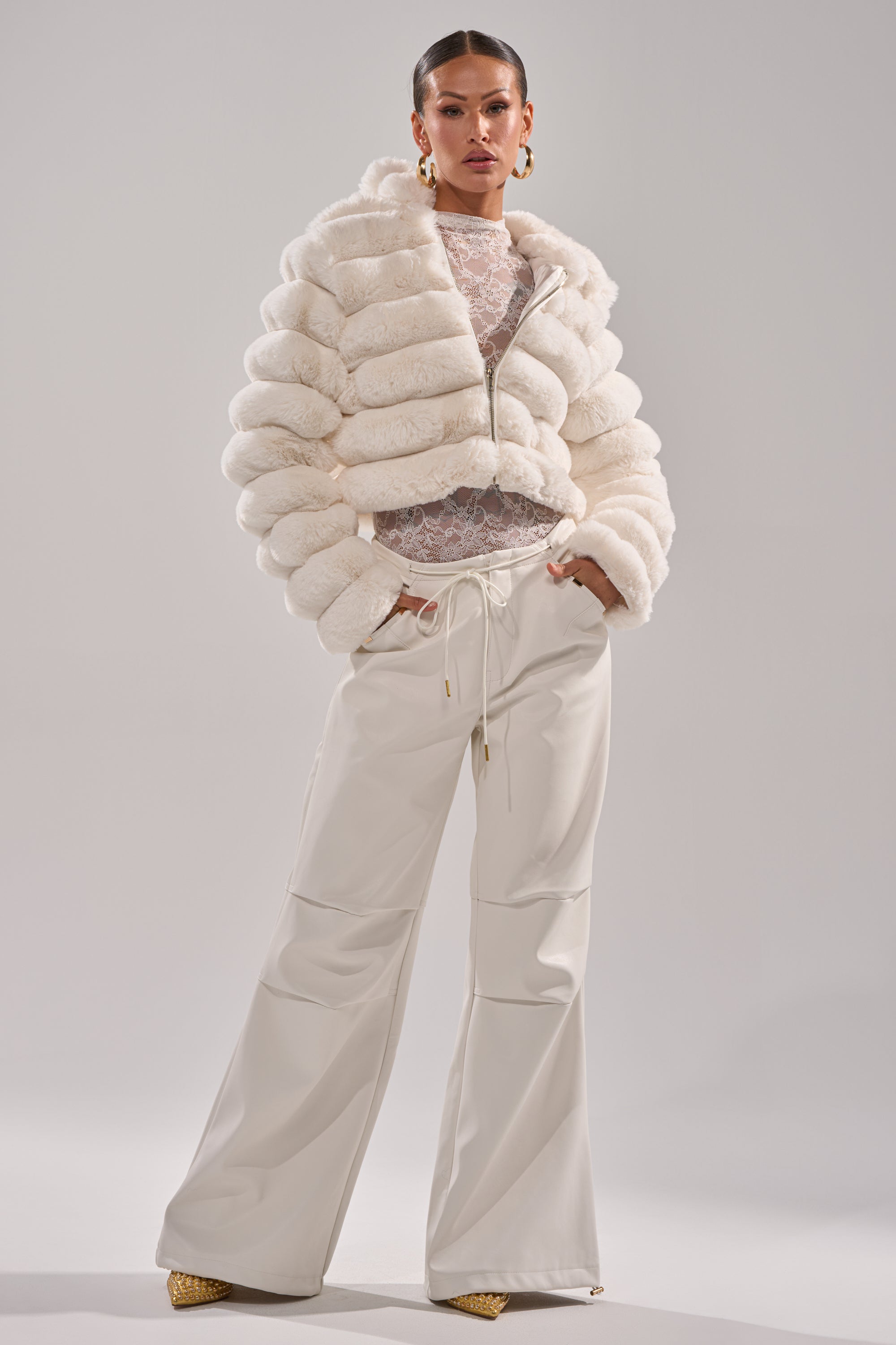 A woman stands confidently against a gray background, wearing the GET WITH IT FAUX LEATHER TROUSER IN WHITE, styled with a white fur jacket, sheer lace top, and gold heels. 6