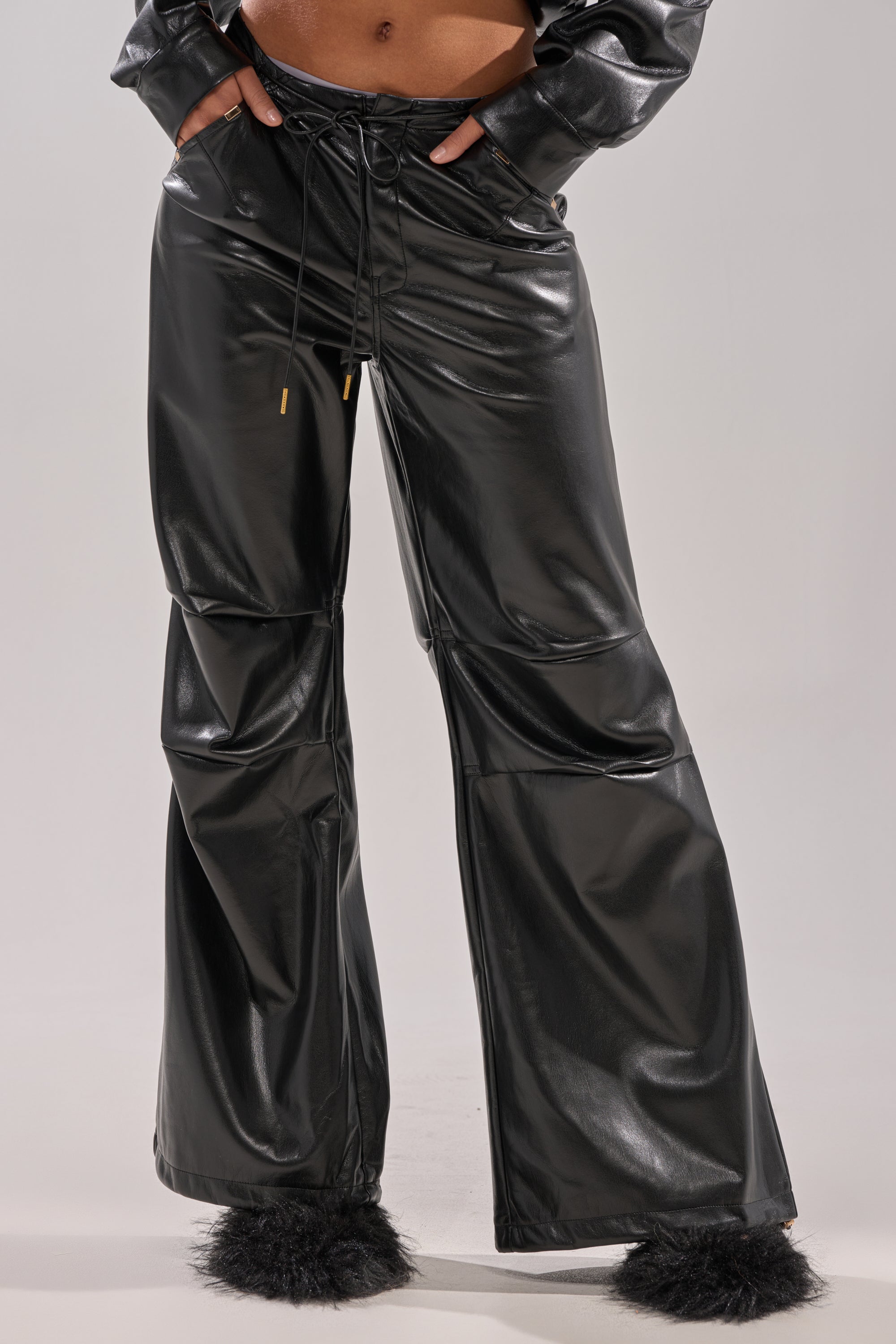 A person wears the GET WITH IT FAUX LEATHER TROUSER—shiny black, high-rise, with drawstring waist and wide-leg fit—paired with black fuzzy slippers. The cropped image shows a plain light background.