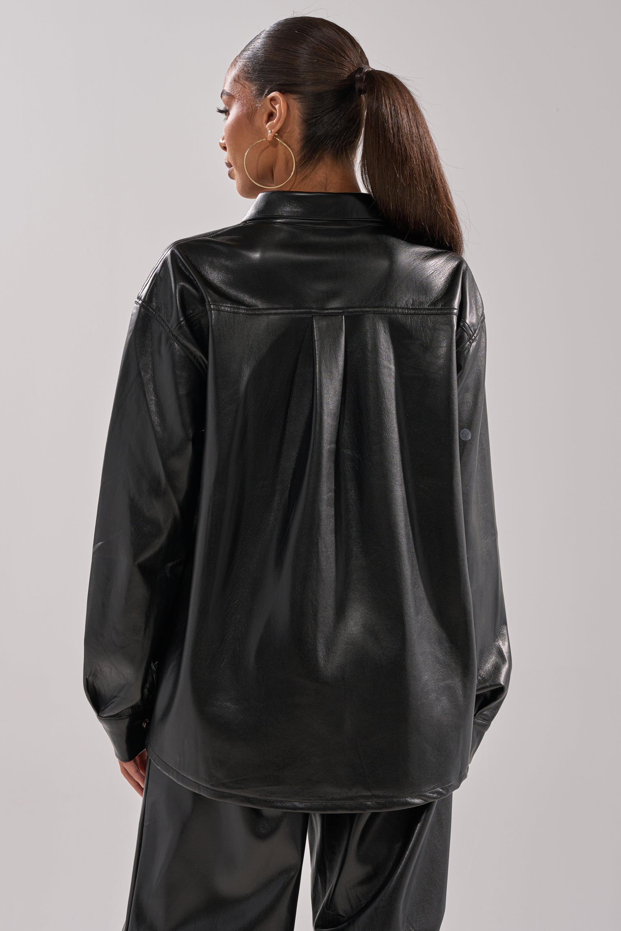 A woman with long brown hair in a ponytail wears the GET WITH IT FAUX LEATHER BUTTON DOWN and matching pants, standing against a plain light gray background with her back to the camera.