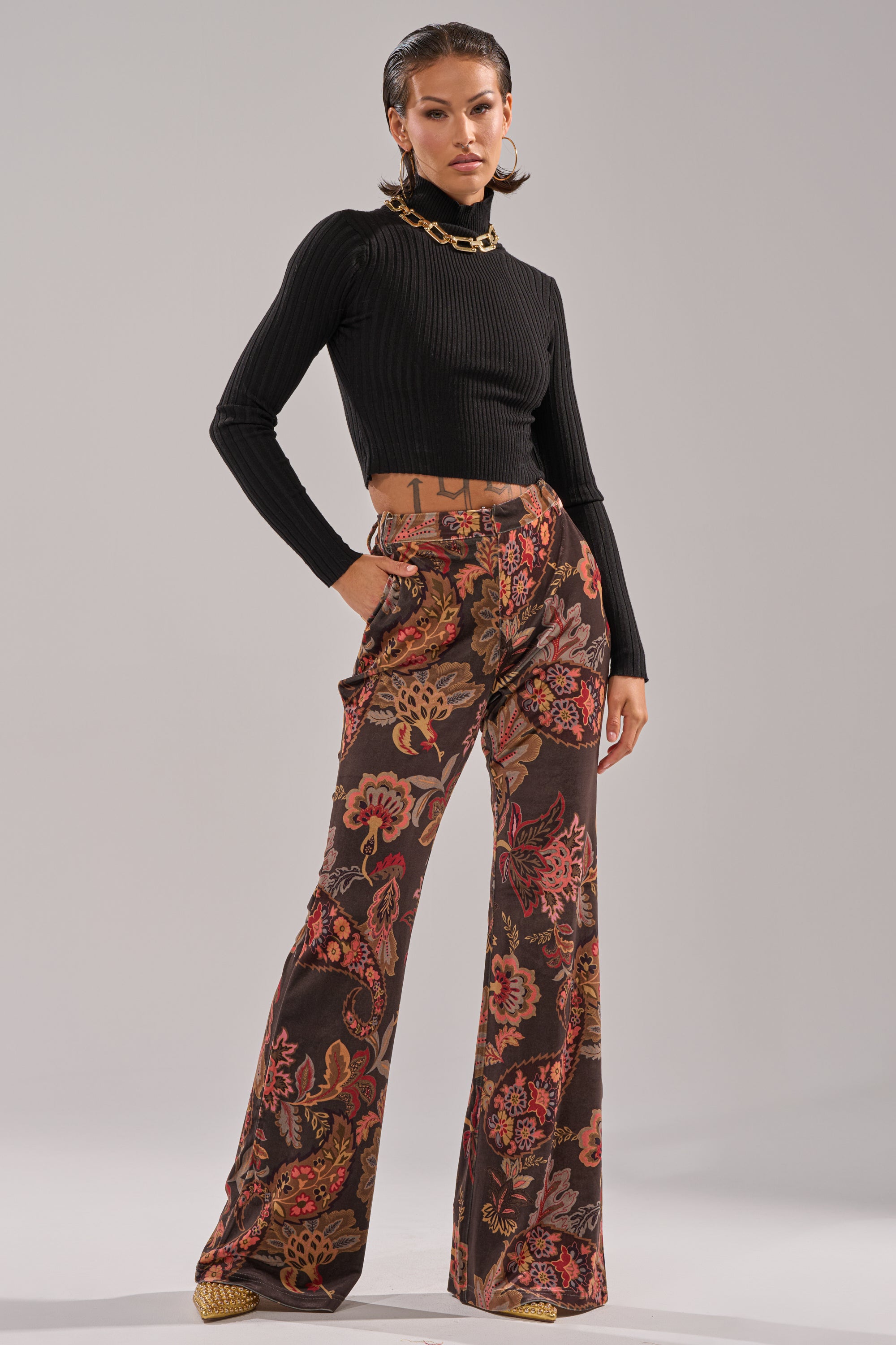 A woman poses confidently in a black ribbed turtleneck and the FALLING IN LOVE FLORAL TROUSER, featuring a bold red, orange, and brown floral pattern. She completes her look with gold jewelry and slicked-back hair.