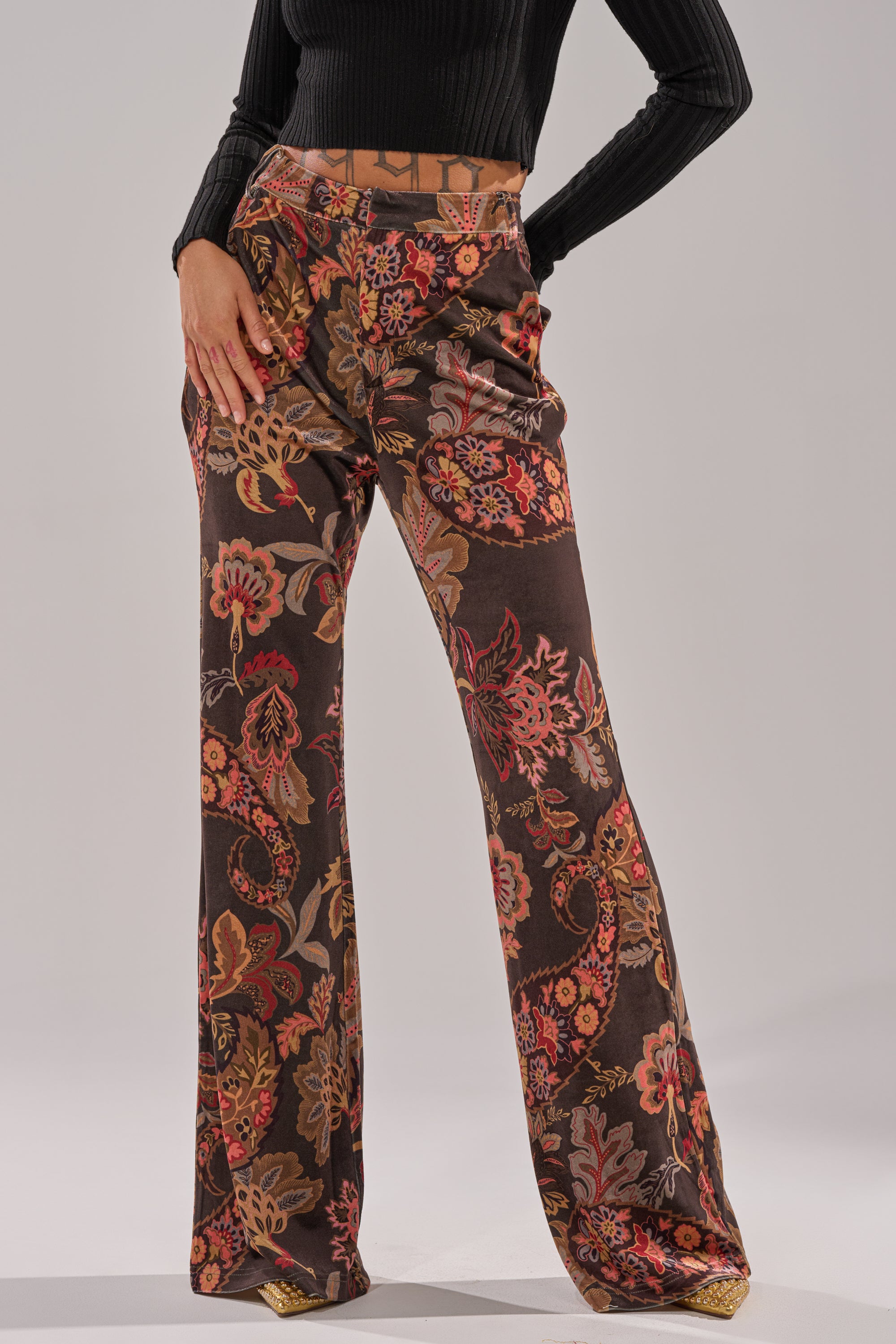 A person wears the FALLING IN LOVE FLORAL TROUSER—high-rise, flared pants with a vibrant red, orange, and yellow floral paisley print—paired with a black ribbed long-sleeve crop top against a plain light background.