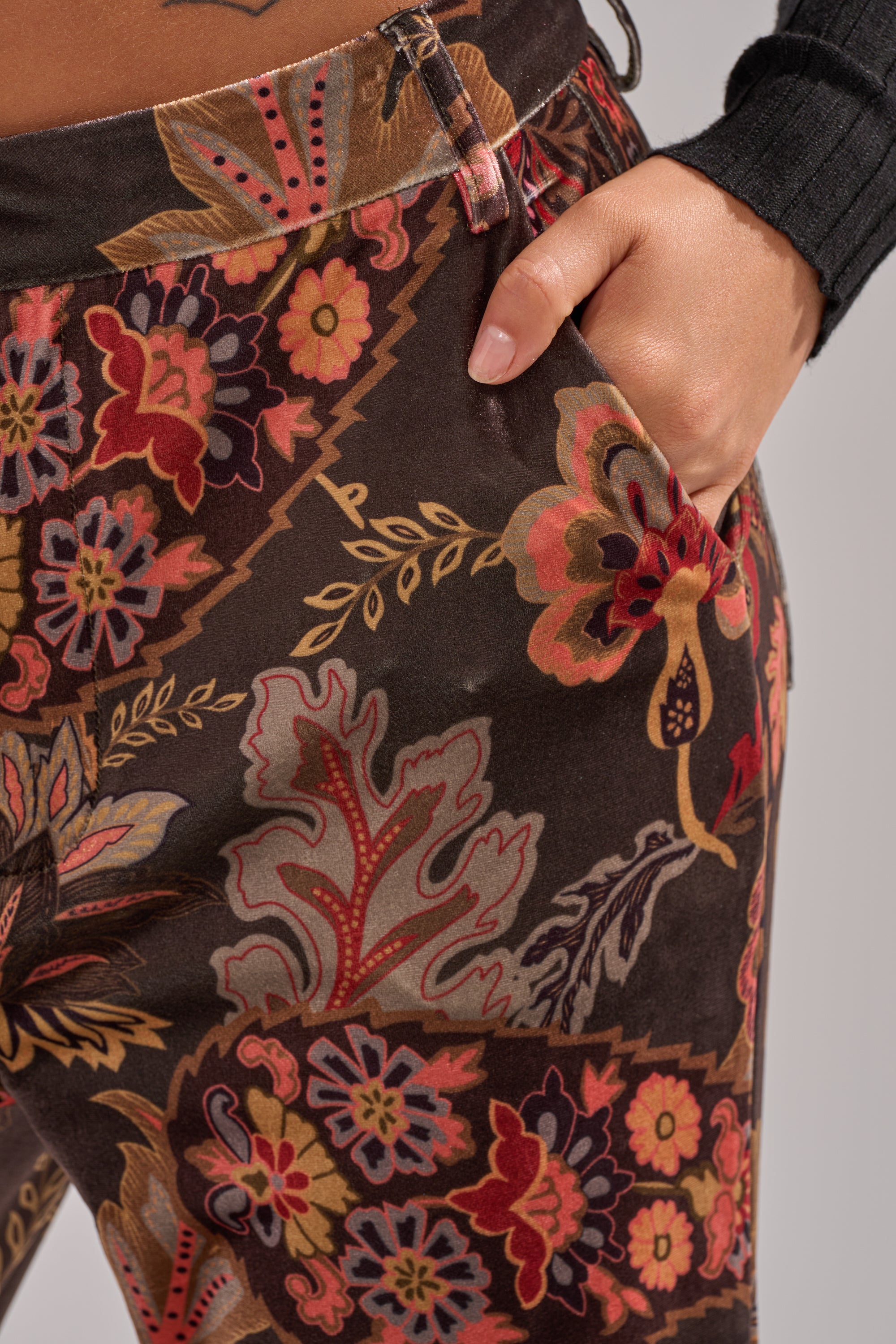 A close-up of a hand in the pocket of the FALLING IN LOVE FLORAL TROUSER, brown high-rise flared pants with a vibrant red, yellow, and orange floral and leaf pattern. The person pairs it with a long-sleeve black top.
