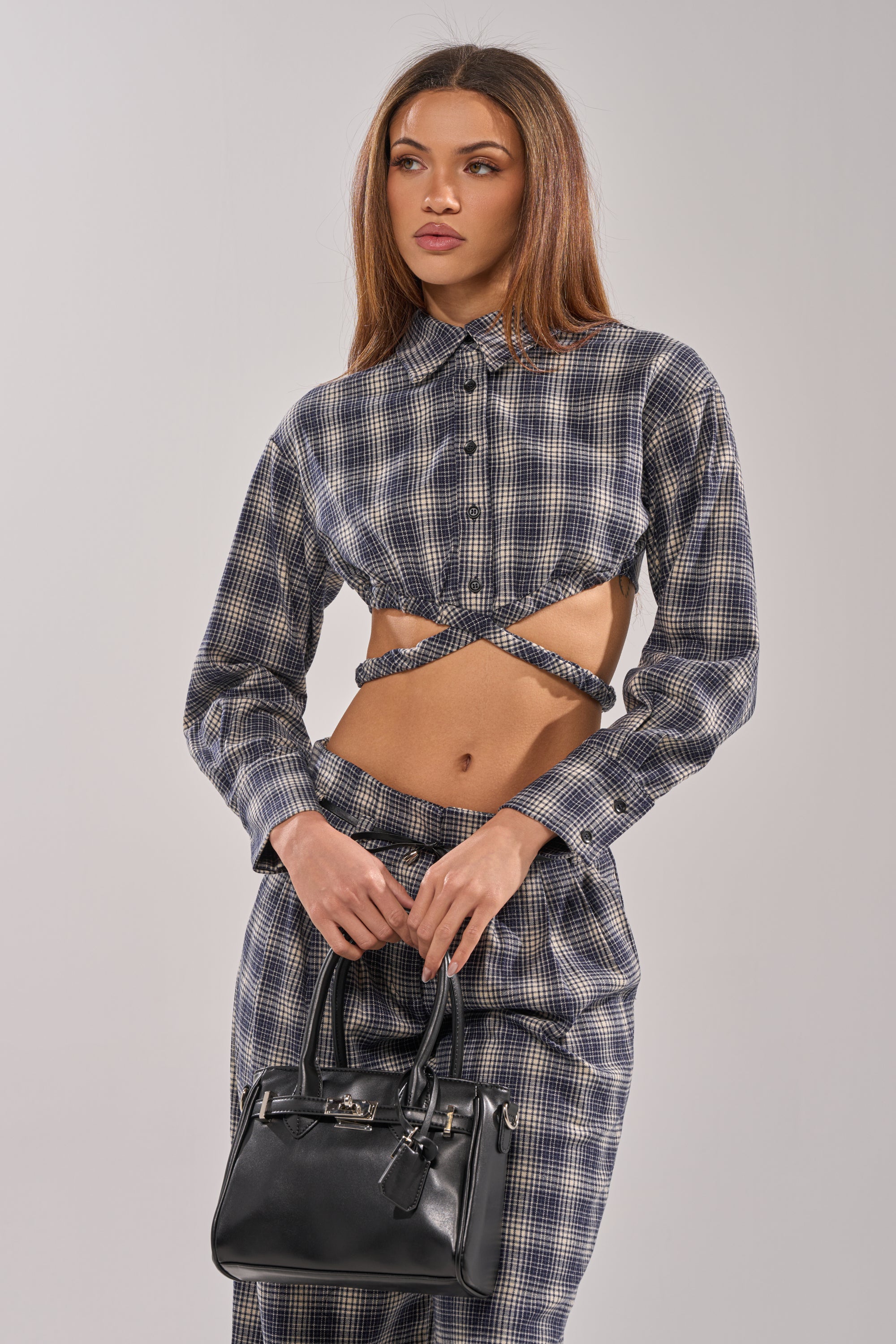 Wearing the JANE PLAID BUTTON DOWN and matching pants, a woman with a confident expression holds a black handbag against a plain light background.