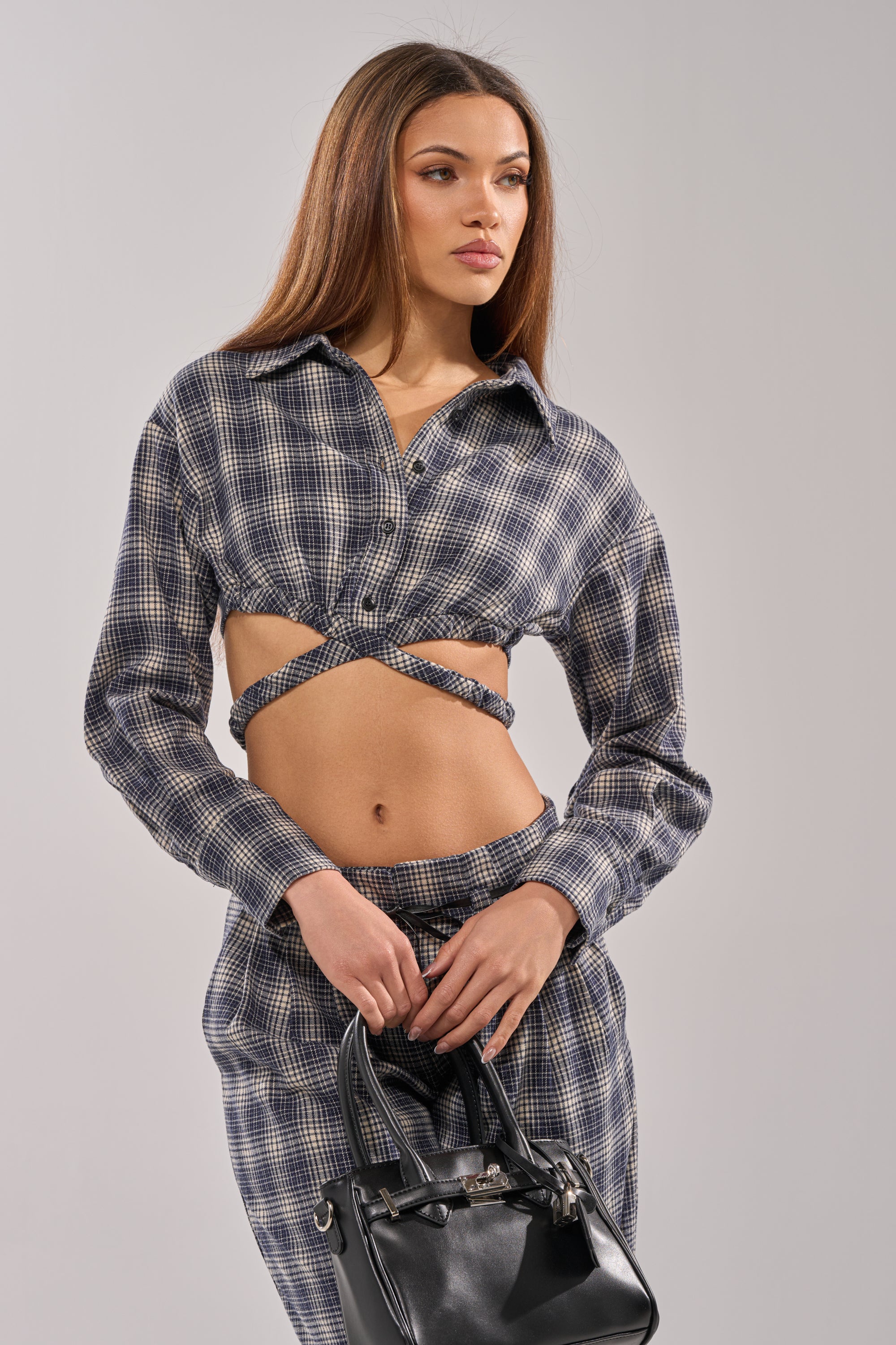 A woman with long brown hair poses in the JANE PLAID BUTTON DOWN, styled as a cropped shirt with matching pants. She holds a small black handbag and looks to the side against a plain light background.