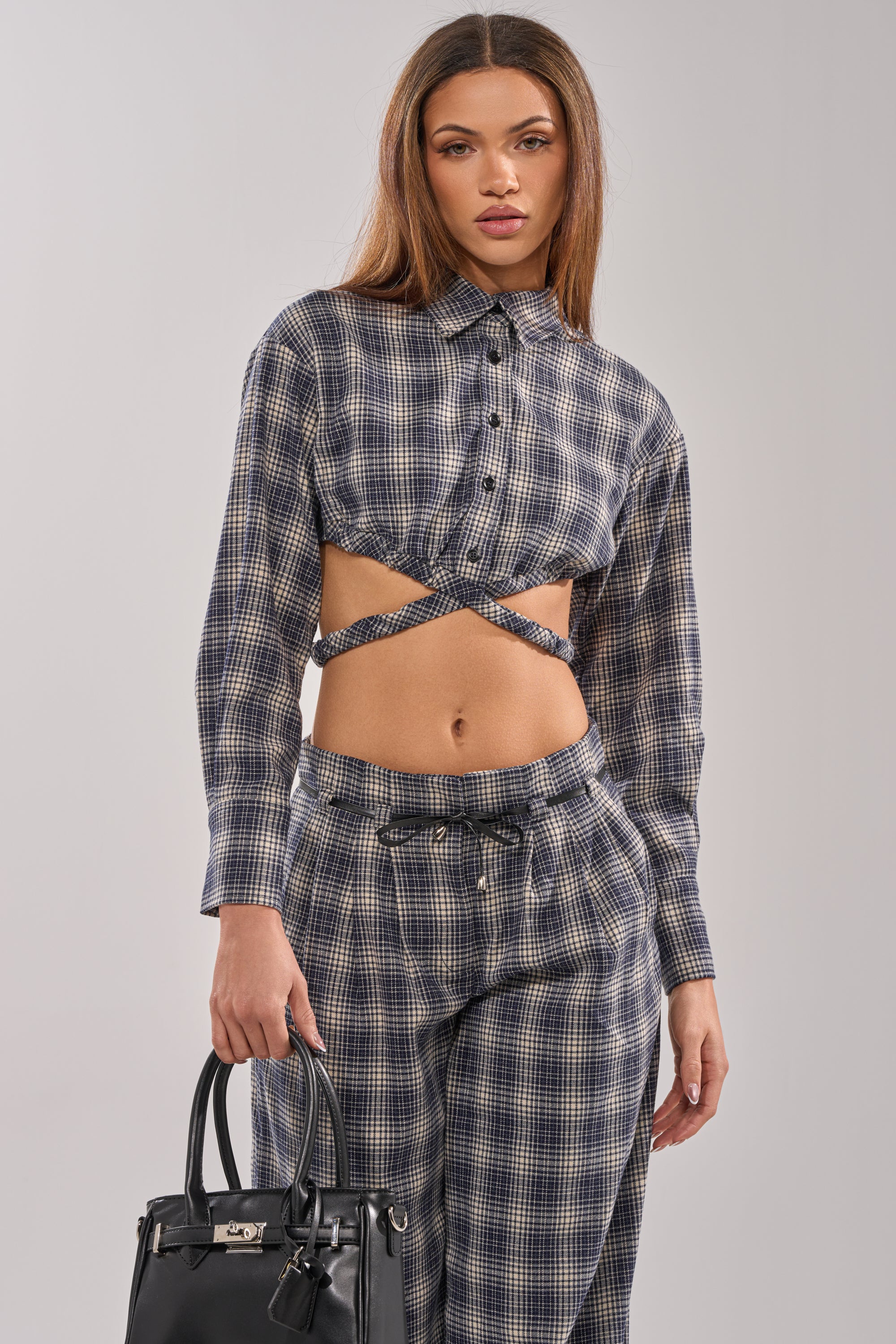 A woman stands against a plain background, wearing the JANE PLAID BUTTON DOWN two-piece outfit with blue and gray plaid high-waisted pants and holding a black handbag.