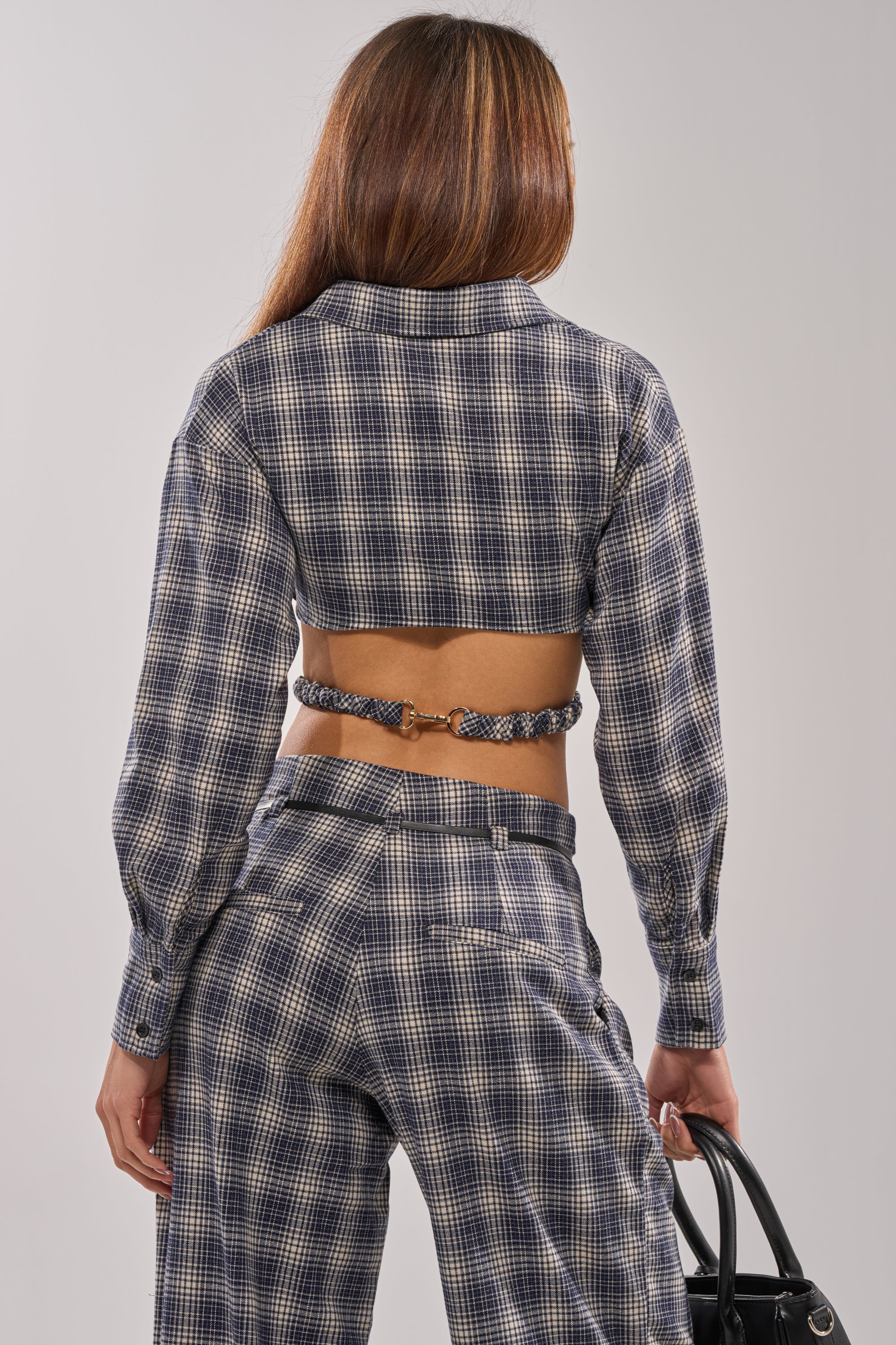 A woman faces away, wearing the JANE PLAID BUTTON DOWN styled open with matching high-waisted plaid pants for a modern streetwear look. She carries a black handbag in her left hand.