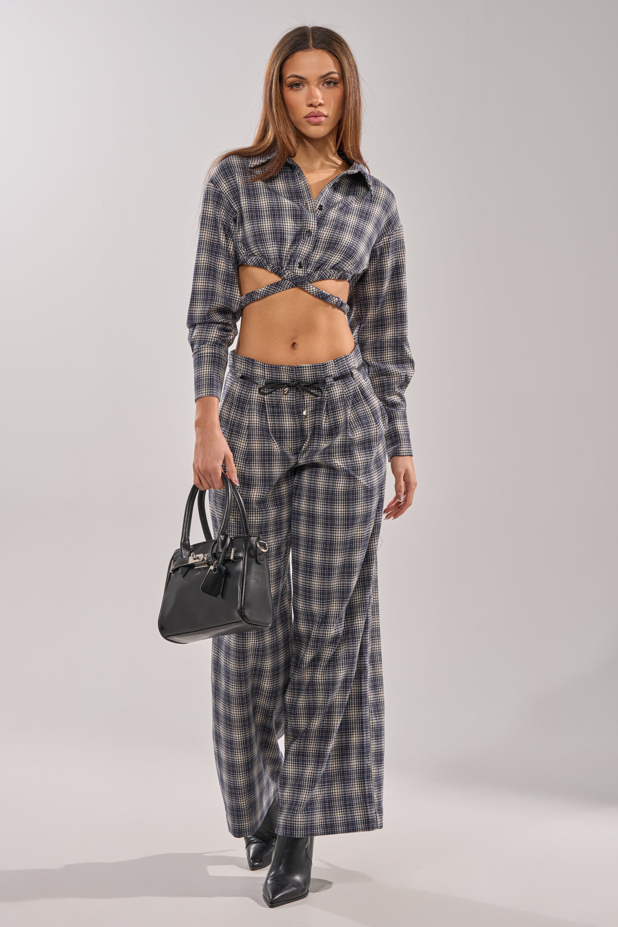 A woman walks forward carrying a black handbag, wearing the JANE PLAID BUTTON DOWN as a cropped top with wide-leg pants and black ankle boots.