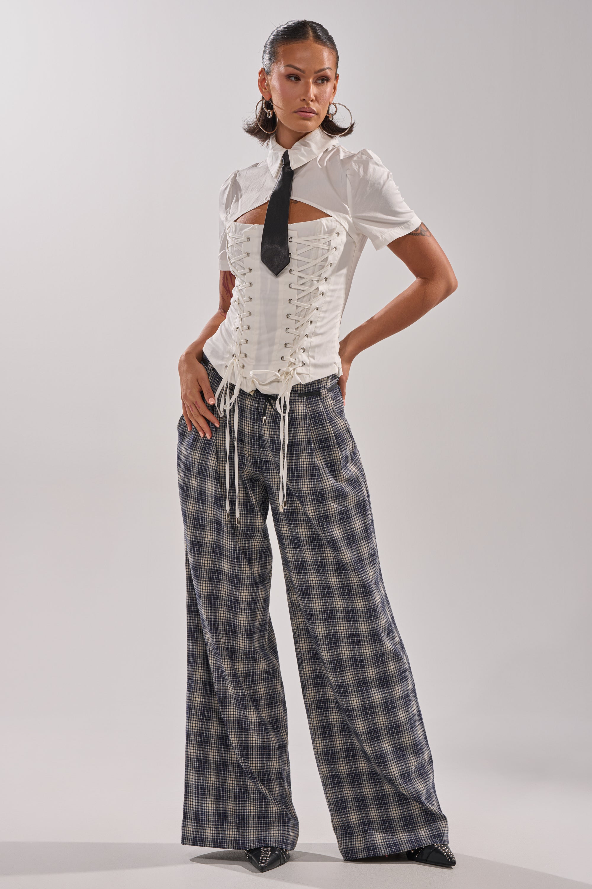 A woman stands against a plain background in JANE PLAID TROUSER, paired with black shoes and a short-sleeve white shirt with a black tie under a white lace-up corset. 1