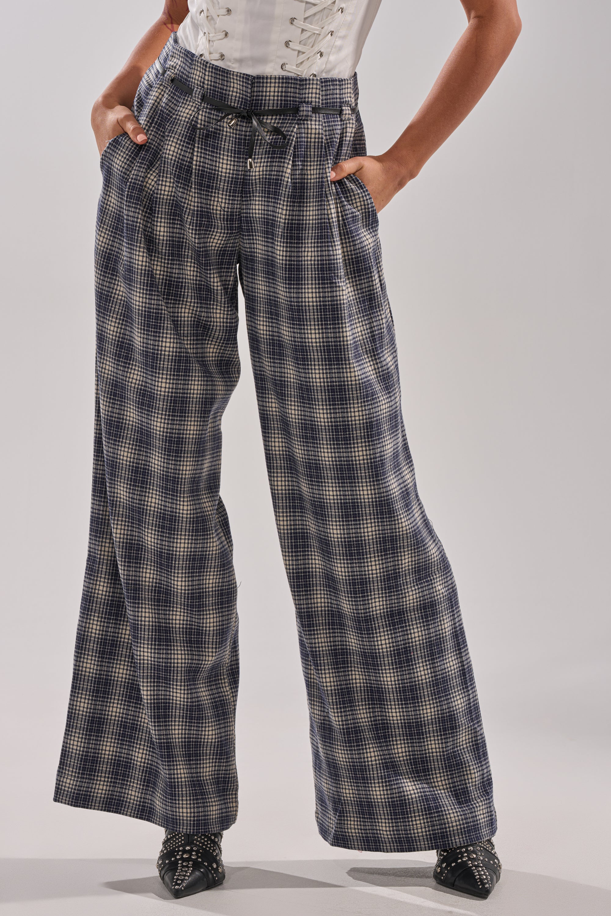 A person stands with hands in pockets against a plain background, wearing the JANE PLAID TROUSER—high-rise, wide-leg with a belted waist—paired with a white corset top and black pointed shoes. 0