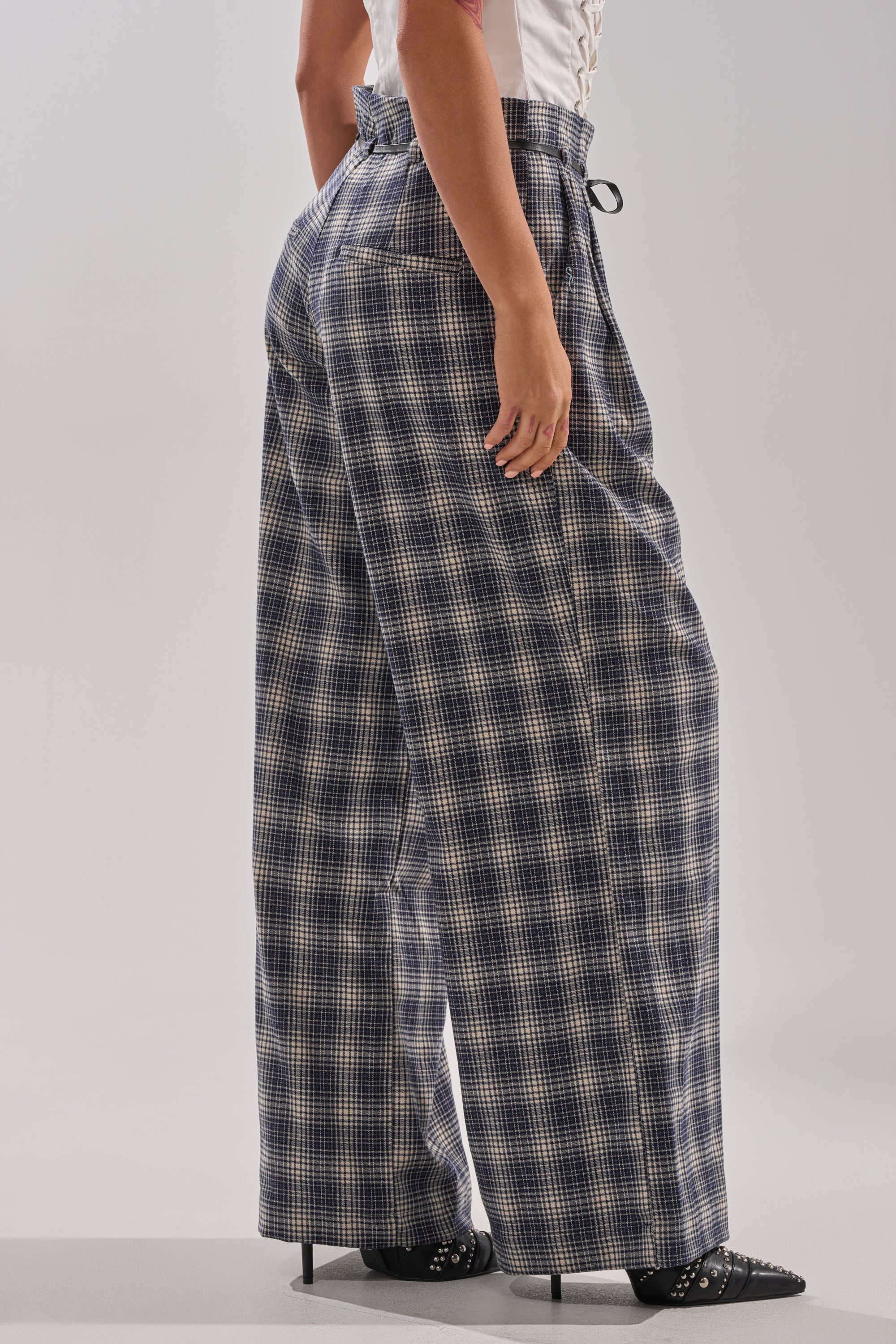 A person stands sideways wearing the JANE PLAID TROUSER, a high-rise wide-leg style in blue and beige plaid, paired with a white top and black heeled shoes against a plain, light background. 3
