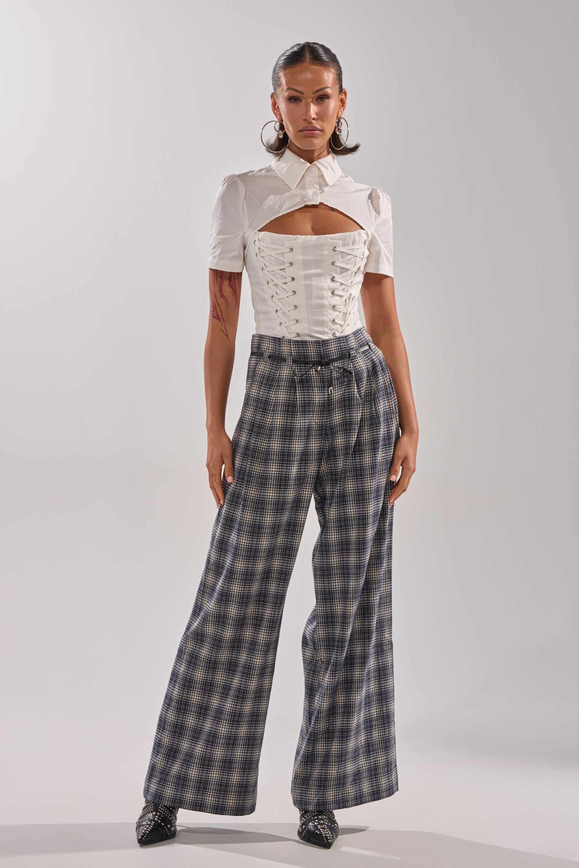 A woman stands in a studio wearing a white corset-style top and the JANE PLAID TROUSER, accessorized with large hoop earrings and black pointed shoes. 5