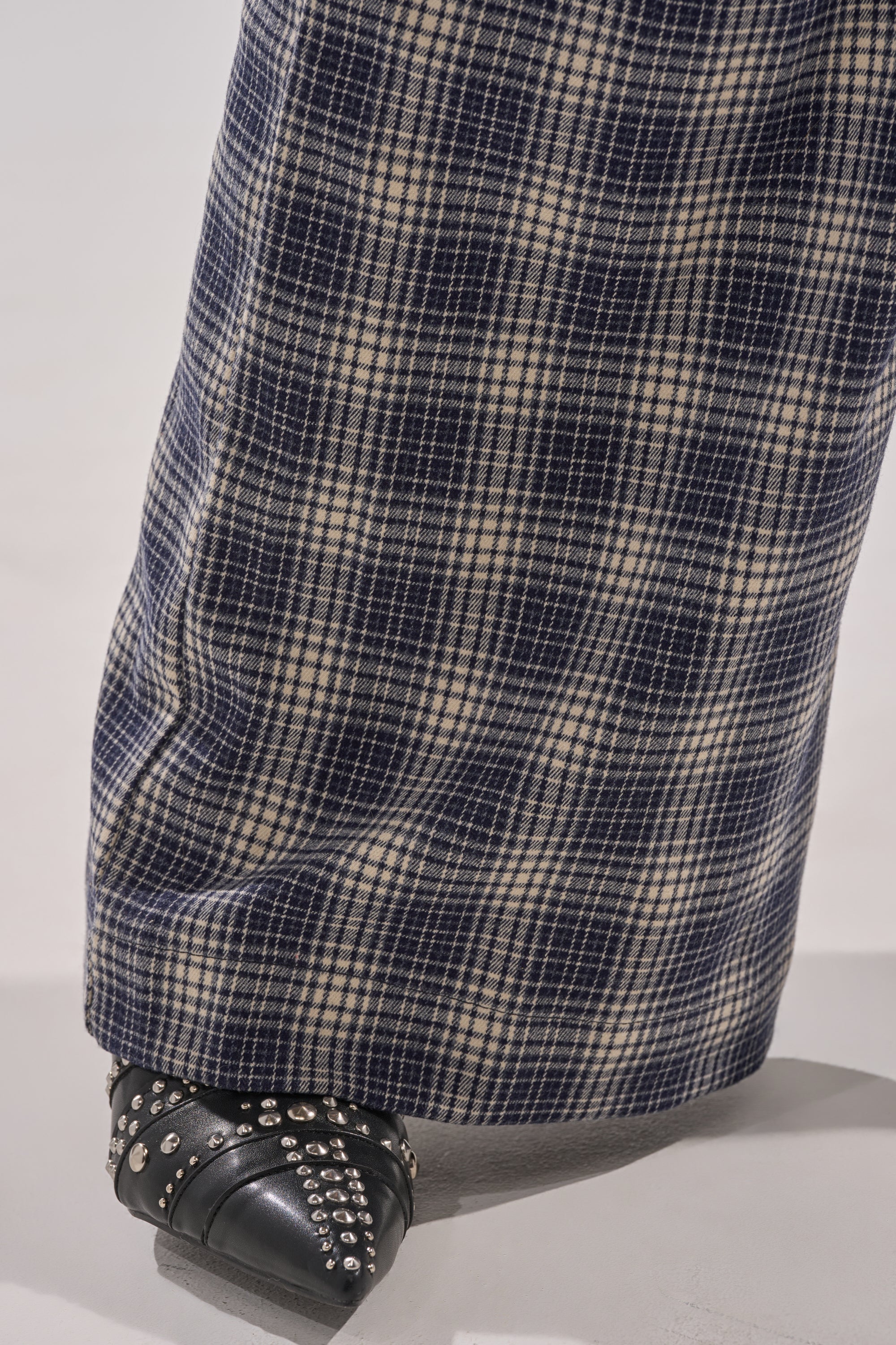 A close-up of a person’s lower leg wearing the JANE PLAID TROUSER in blue and white plaid, paired with a black studded shoe. The high-rise, wide-leg pants partially cover the stylish footwear. 8