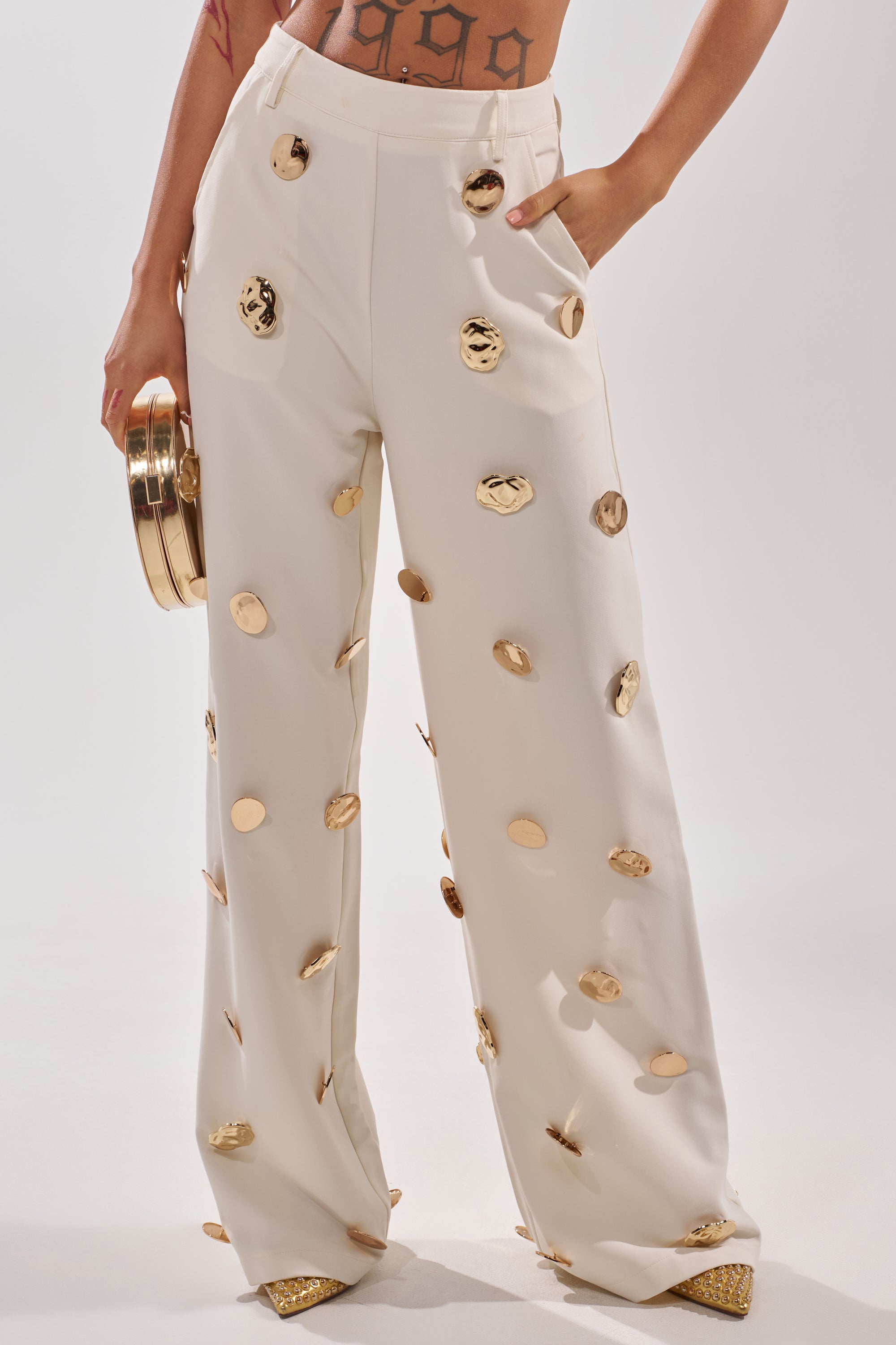 Wearing the SARASOTA EMBELLISHED TROUSER, high-waisted and wide-leg with bold gold medallions, and holding a matching gold circular clutch. Only the lower torso and legs are visible. 1