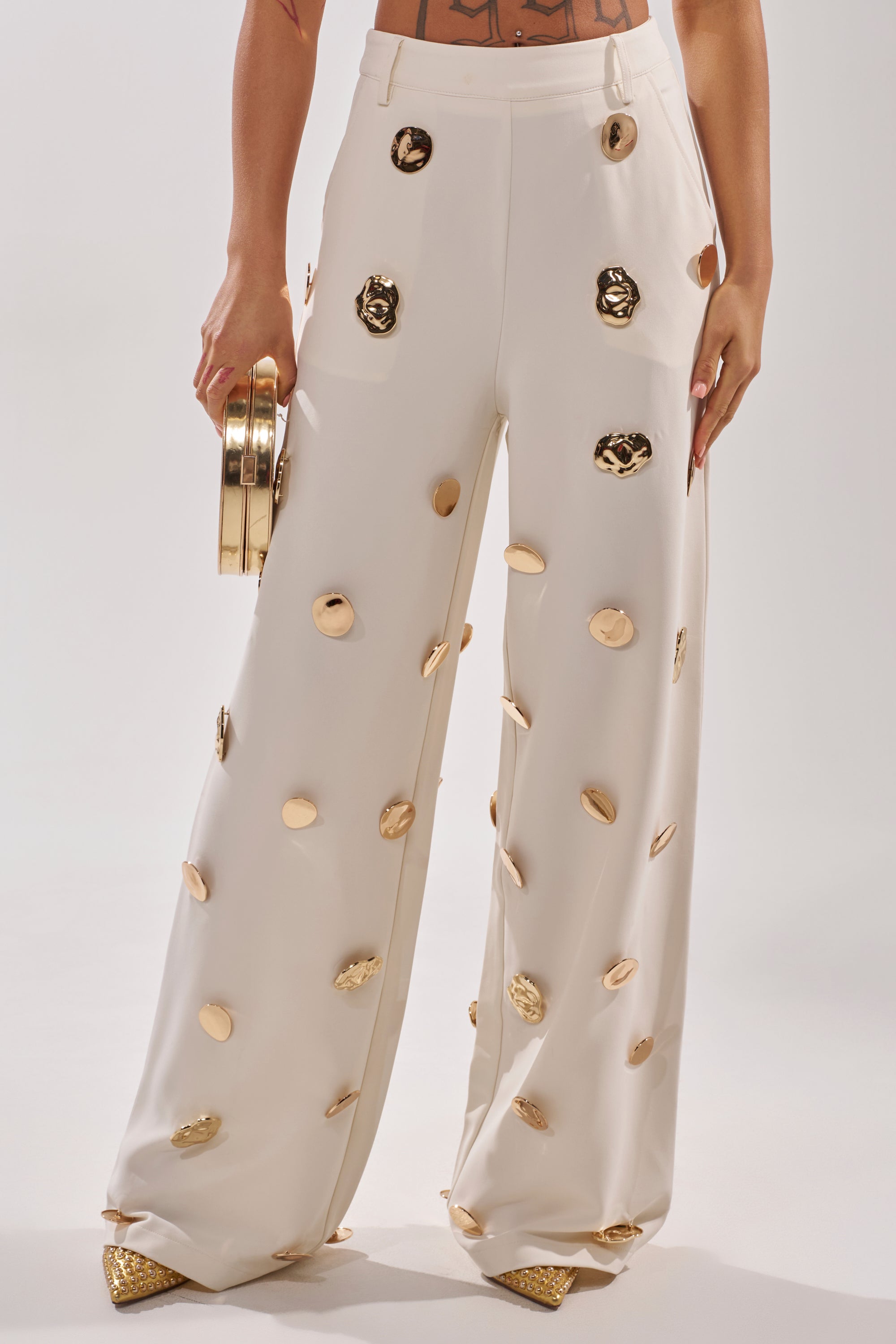 Wearing the SARASOTA EMBELLISHED TROUSER with gold medallion accents, a person holds a round gold clutch and wears pointed gold shoes against a plain, light background. 2