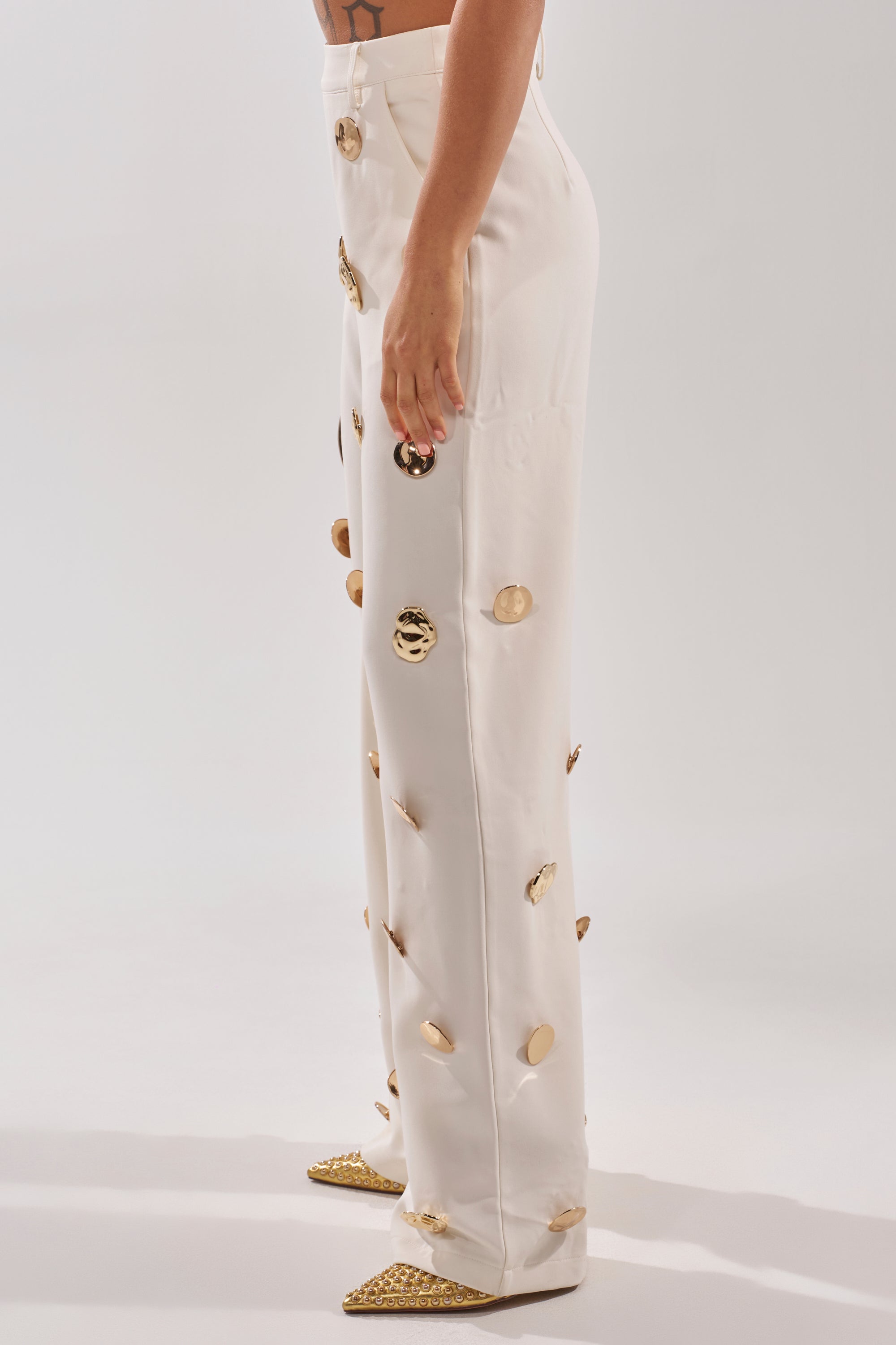A person stands sideways in the SARASOTA EMBELLISHED TROUSER, featuring high-waisted wide legs and gold medallion details, paired with pointed gold shoes against a plain light background. 3