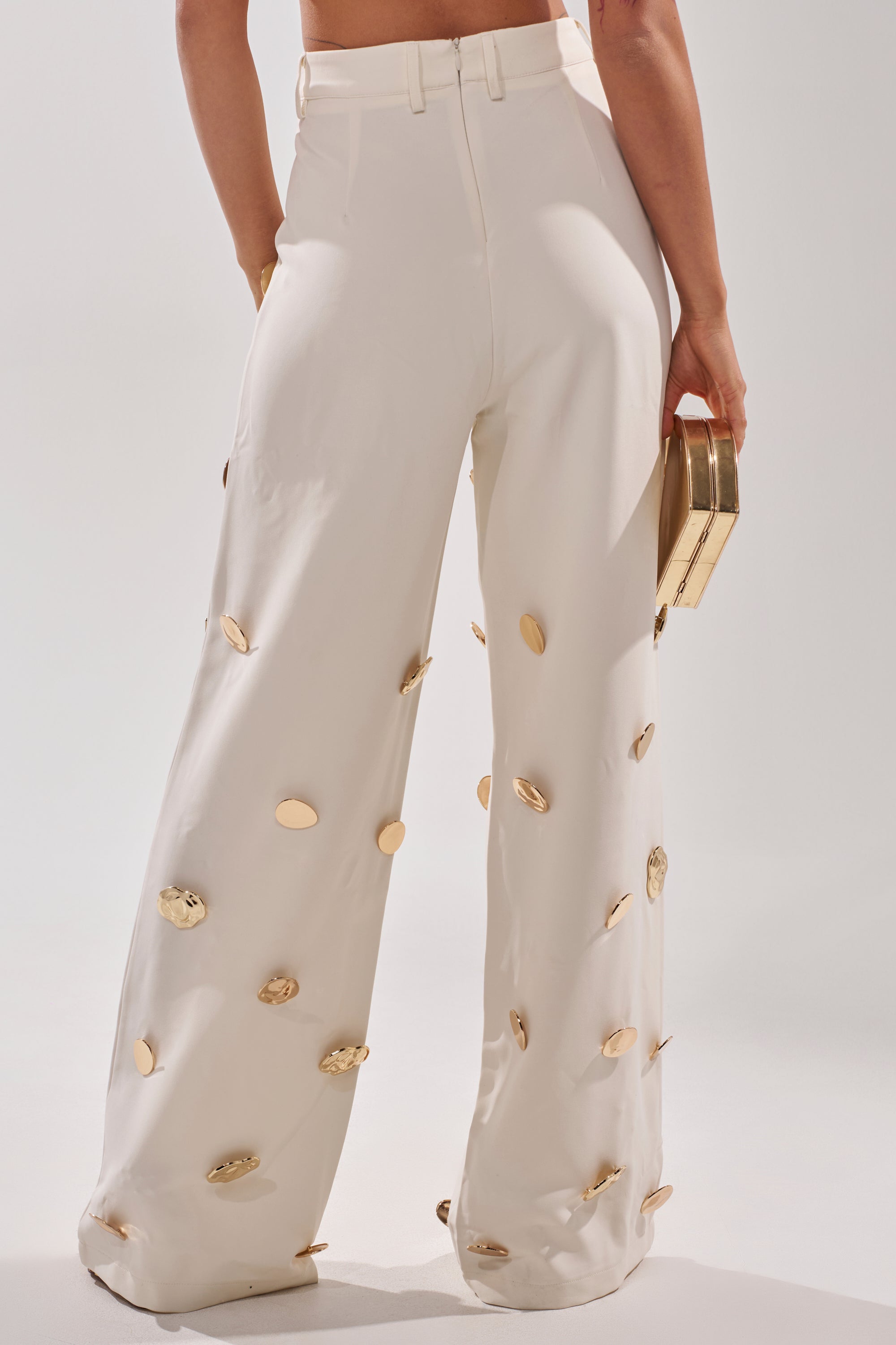 A person faces away, wearing the SARASOTA EMBELLISHED TROUSER—high-rise white wide-leg pants with large gold circular accents—and holds a small gold box clutch against a plain light background. 4
