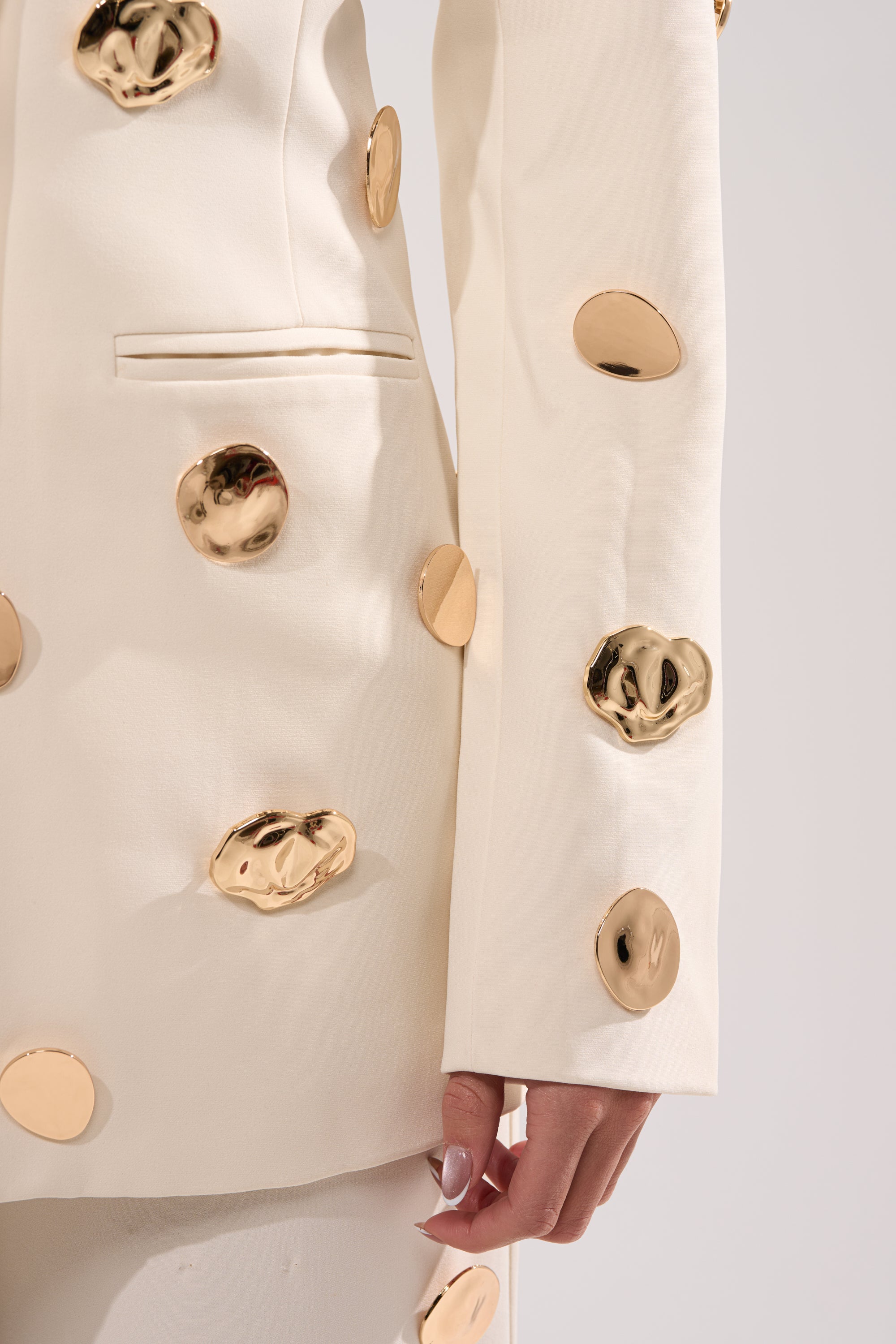 A close-up of the SARASOTA EMBELLISHED BLAZER—a white blazer featuring bold, shiny gold circular embellishments. A manicured hand rests at the side, set against a neutral light background. 9