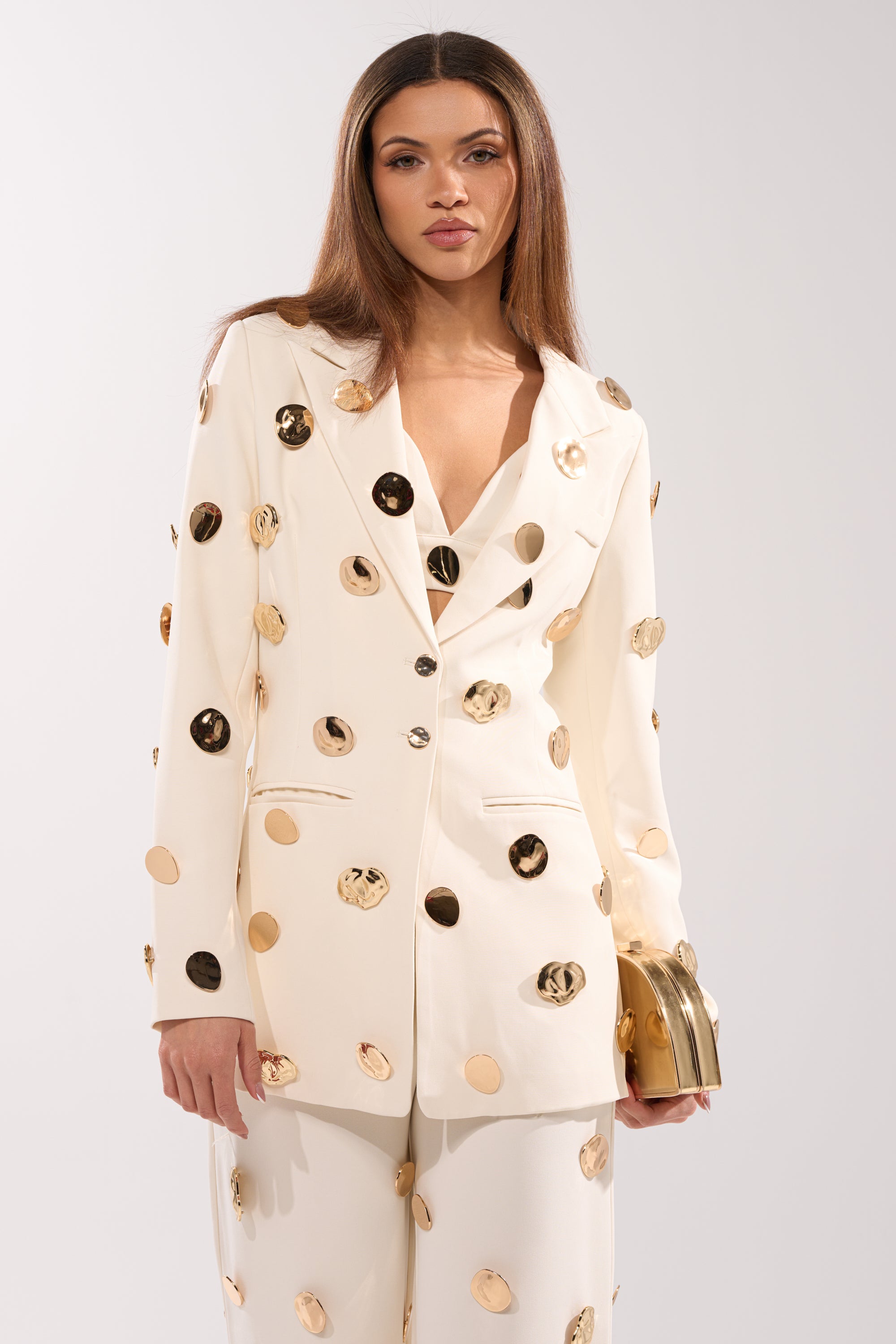 A woman with long straight hair and a neutral expression wears the SARASOTA EMBELLISHED BLAZER, a white piece detailed with gold discs, and holds a small gold clutch against a plain light background. 2