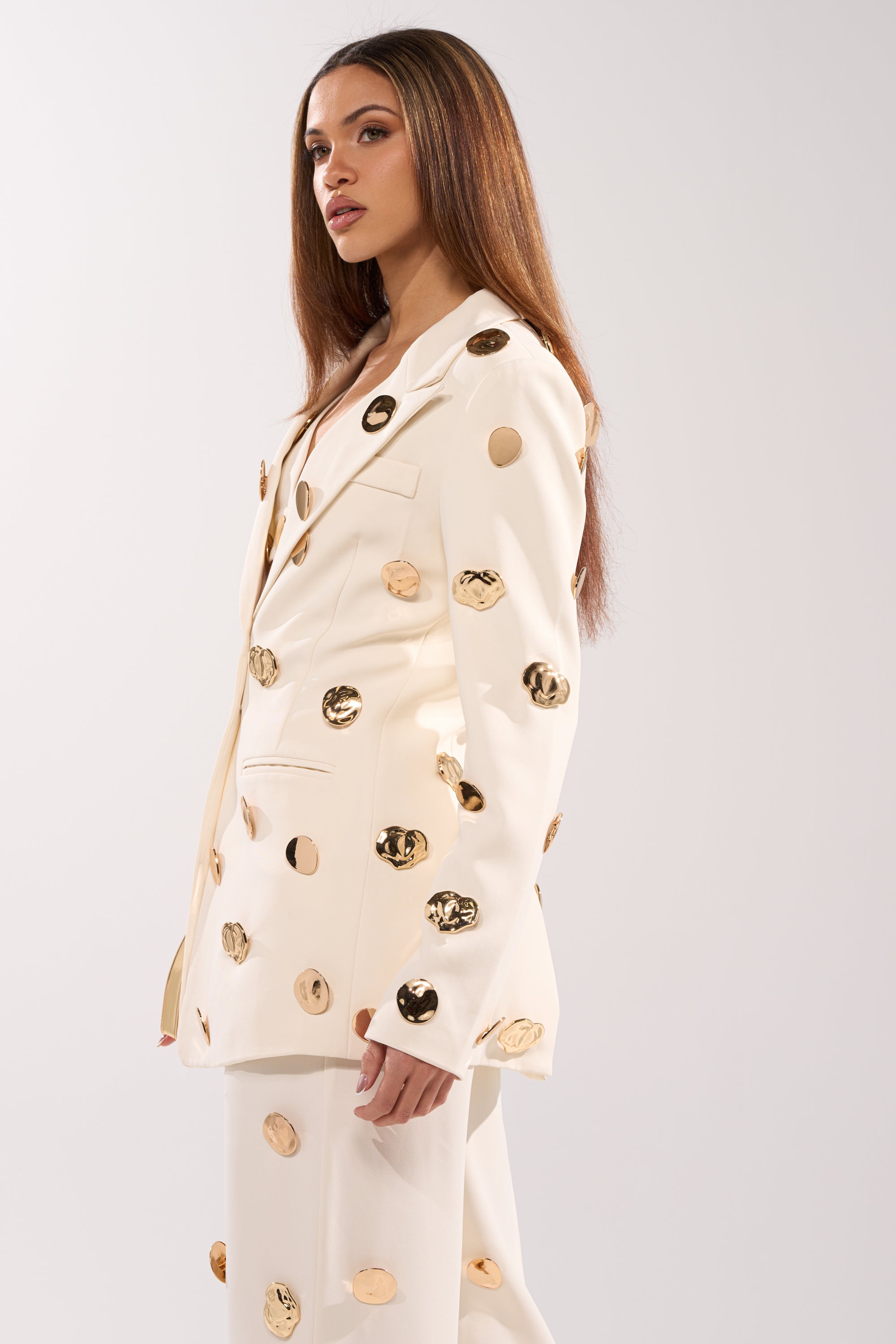 A woman with long straight hair wears the SARASOTA EMBELLISHED BLAZER over a cream suit, standing against a plain light background and gazing slightly to the side. 4