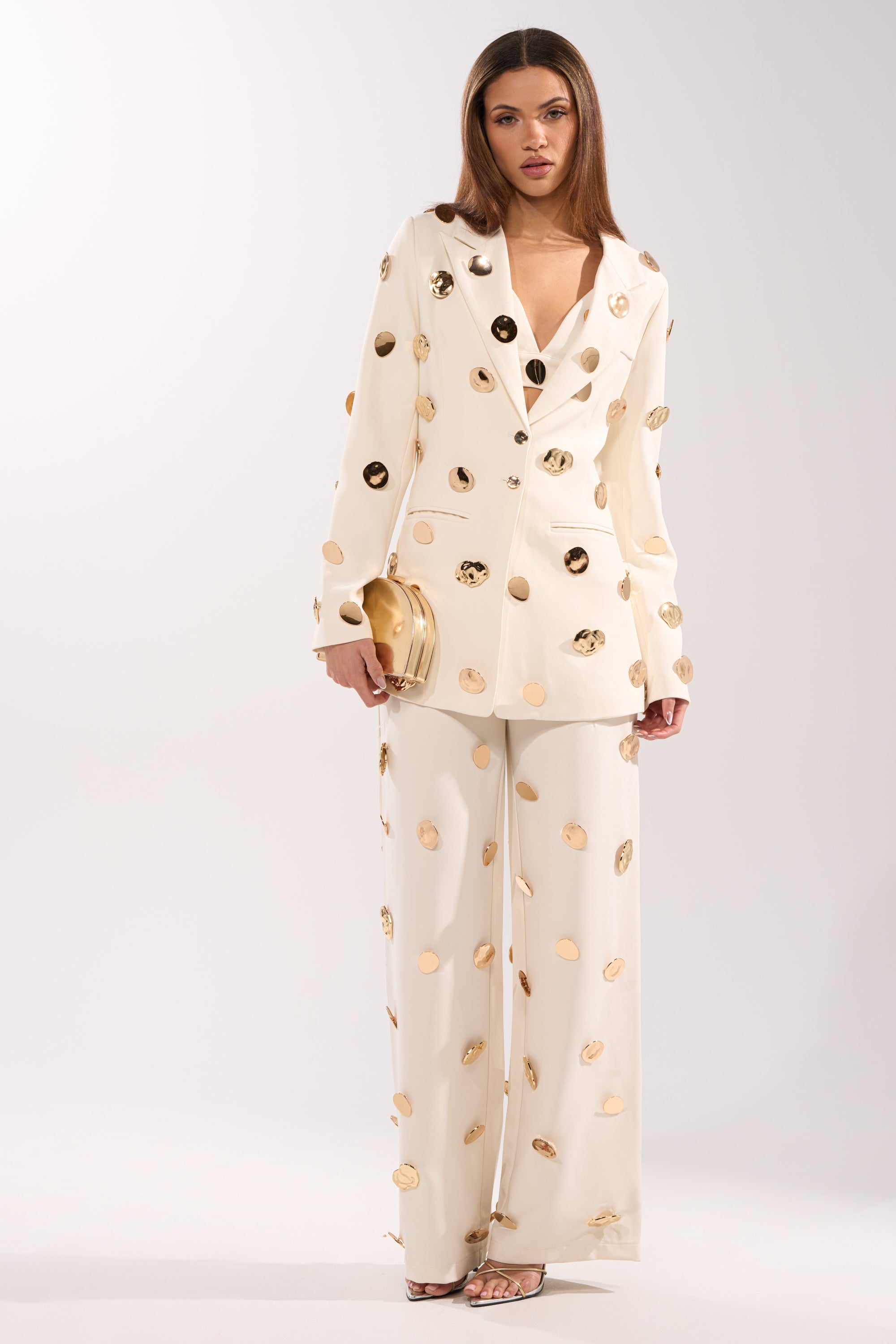 A woman stands against a plain white background wearing the SARASOTA EMBELLISHED BLAZER with matching pants, a gold clutch bag, and open-toe heels. 7