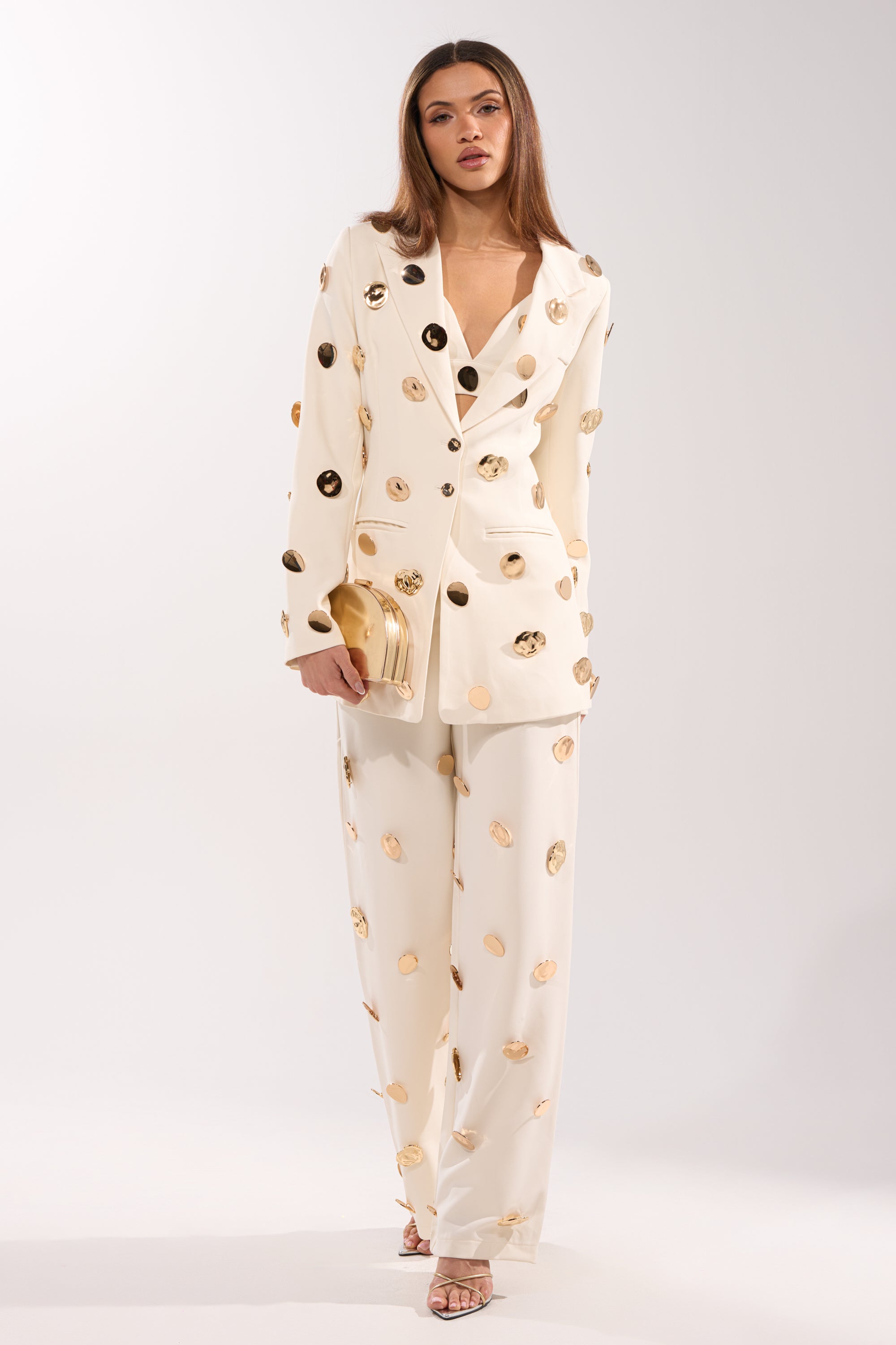 A woman wears the SARASOTA EMBELLISHED BLAZER—cream-colored with large gold polka dots—paired with a gold clutch and heeled sandals, standing against a plain light background. 8