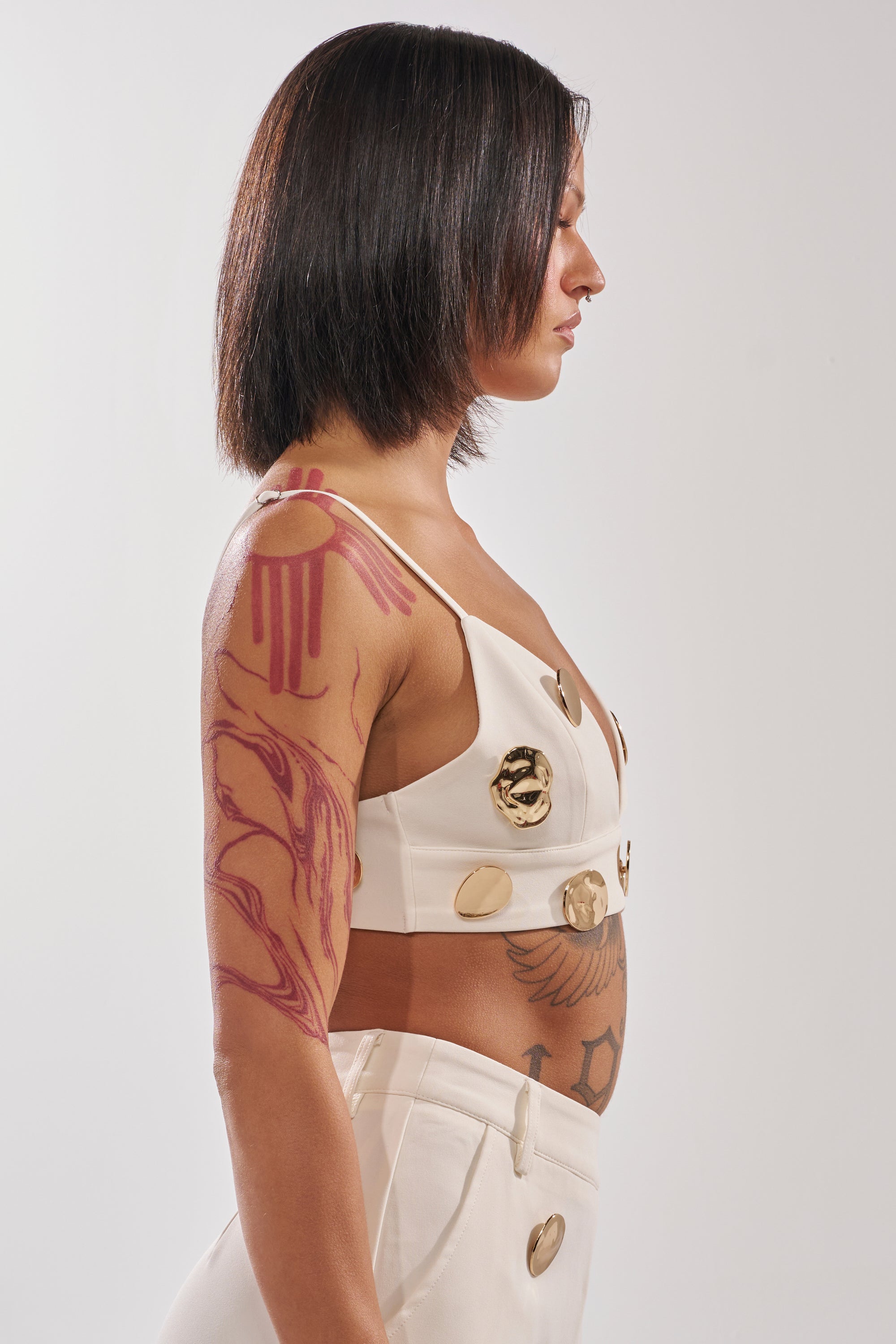 A person with straight, shoulder-length dark hair stands in profile, wearing the SARASOTA EMBELLISHED BRALETTE— a white AKIRA Label piece with gold-tone disc accents—displaying red and black tattoos against a plain background. 3