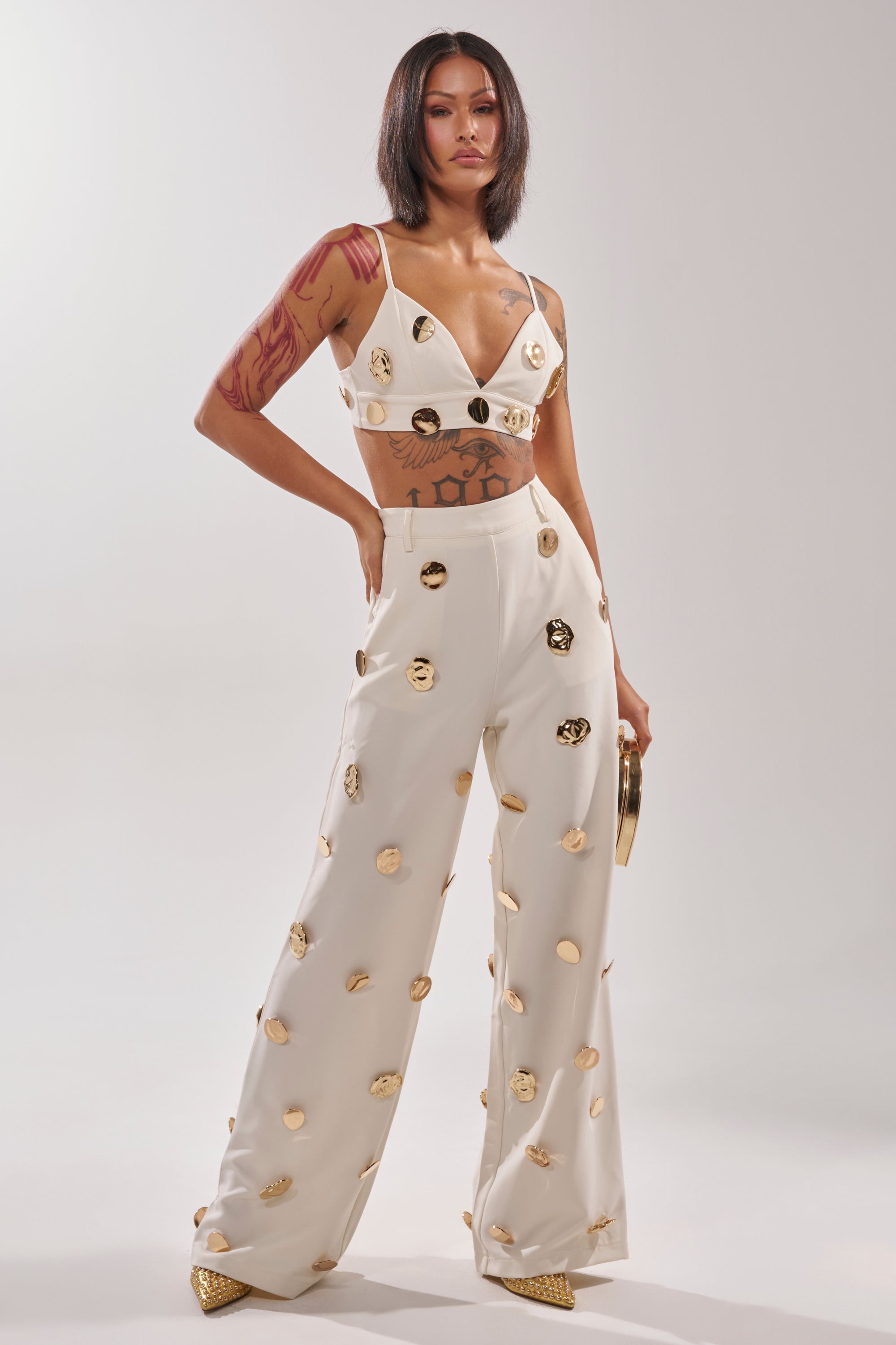A woman with shoulder-length dark hair and visible tattoos models the SARASOTA EMBELLISHED BRALETTE and wide-legged pants decorated with gold-tone discs, paired with pointed gold shoes, standing confidently against a plain background. 5