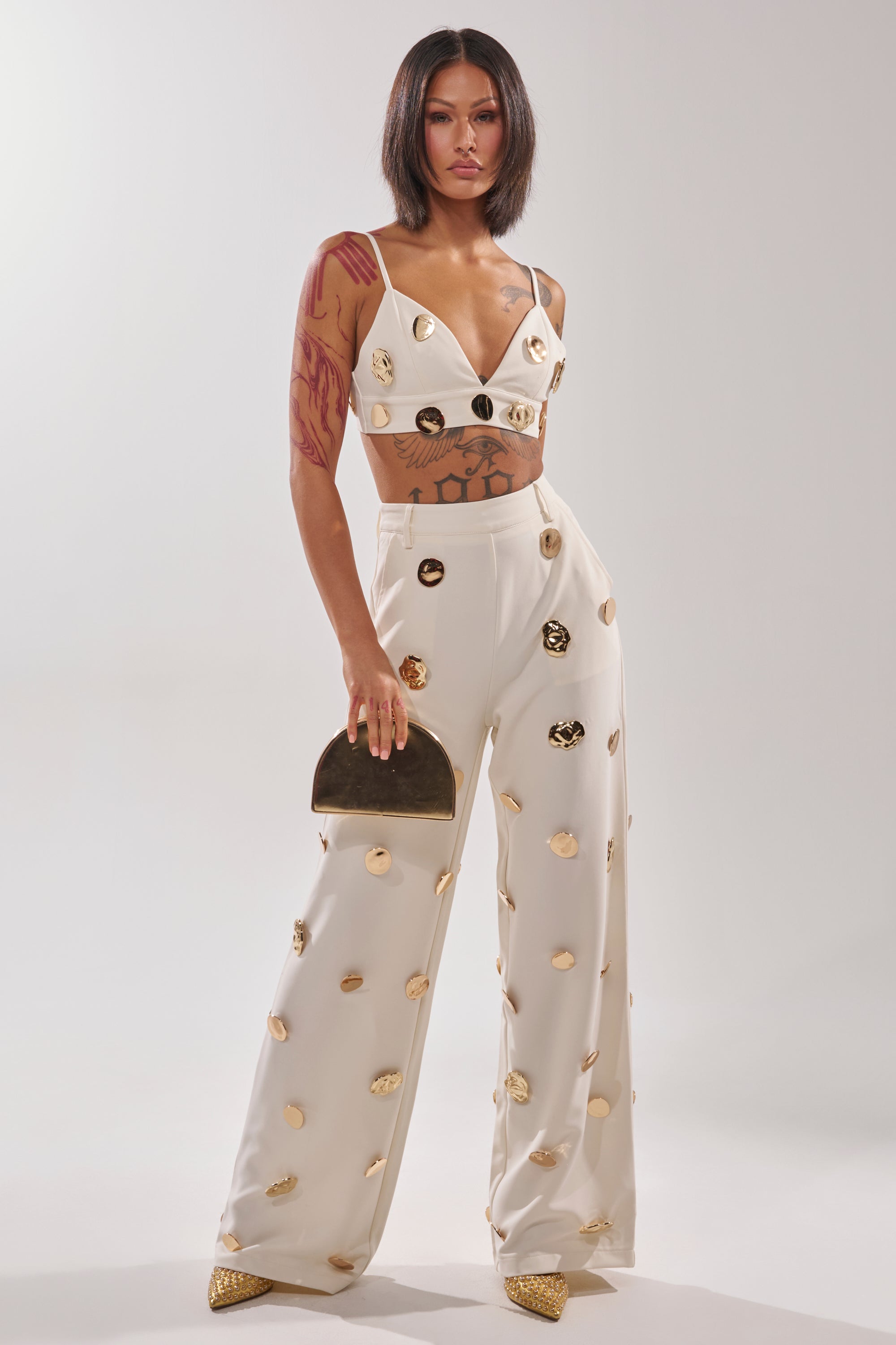 A woman with a sleek bob wears the SARASOTA EMBELLISHED BRALETTE and wide-leg pants with gold-tone disc details. She holds a metallic clutch, sports pointed gold shoes, and poses confidently against a plain background. 6