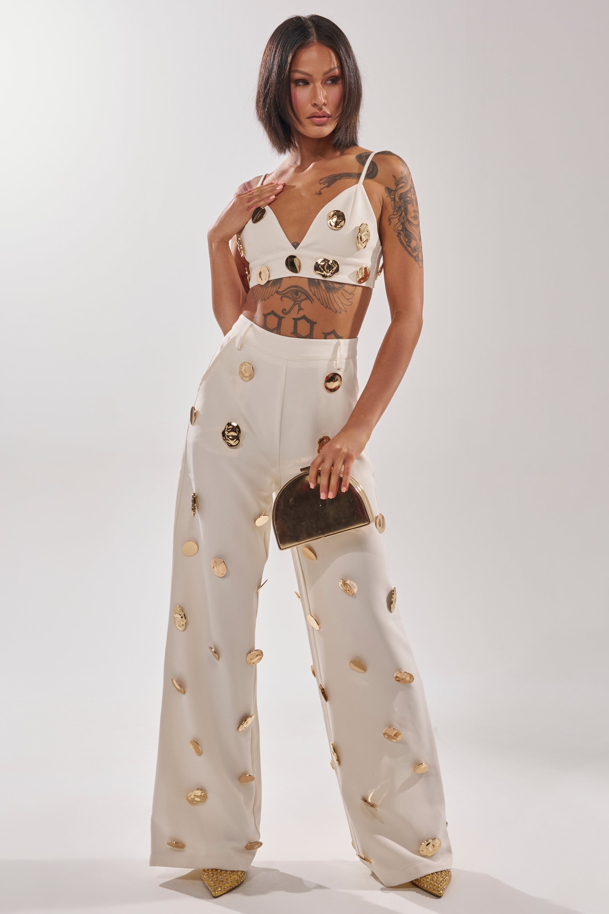 A woman with shoulder-length hair and arm tattoos poses confidently in the SARASOTA EMBELLISHED BRALETTE, paired with high-waisted pants featuring gold-tone disc details, a metallic gold clutch, and gold pointed shoes. 7